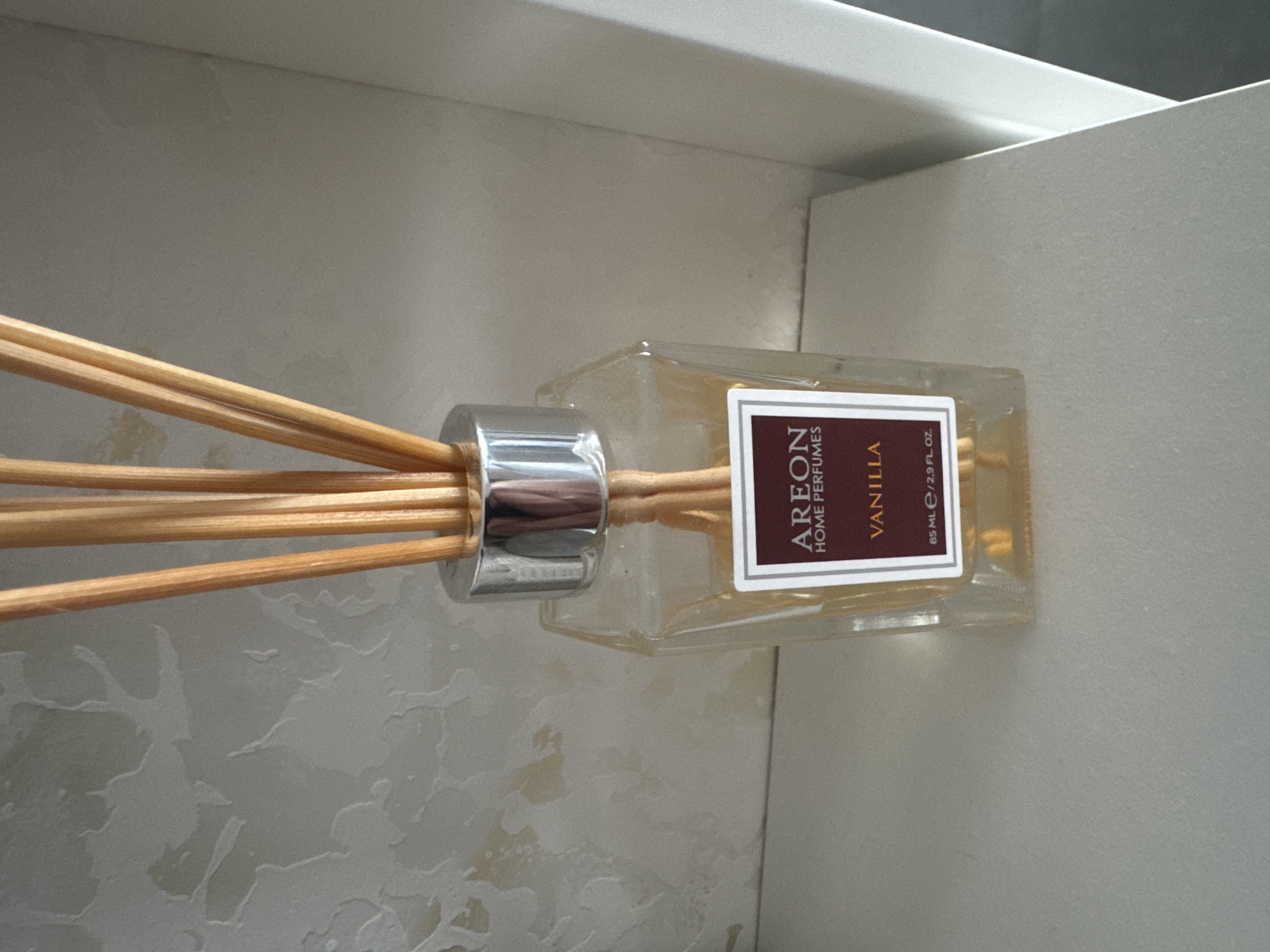This is a close-up, eye-level shot of an "Areon Home Perfumes" reed diffuser with a vanilla scent, placed in what appears to be a bathroom or cabinet setting. The diffuser has a clear glass bottle containing a yellowish liquid, topped with a silver metal cap. Several thin wooden reeds are inserted into the cap, protruding upwards and outwards. A rectangular label on the bottle reads "AREON HOME PERFUMES" and "VANILLA" in a dark red and white font. Below "VANILLA", smaller text indicates "85 ML / 2.9 FL. OZ.". The background is a textured, light-colored surface that appears to be tiled or wallpapered, with some subtle light and dark shading. The diffuser is positioned horizontally, with the reeds extending to the left and the bottle to the right. The setting is indoors, with no indication of the time of day or weather. The overall impression is of a staged product photograph or a casual snapshot of an item in its intended environment. There are no people visible in the image.