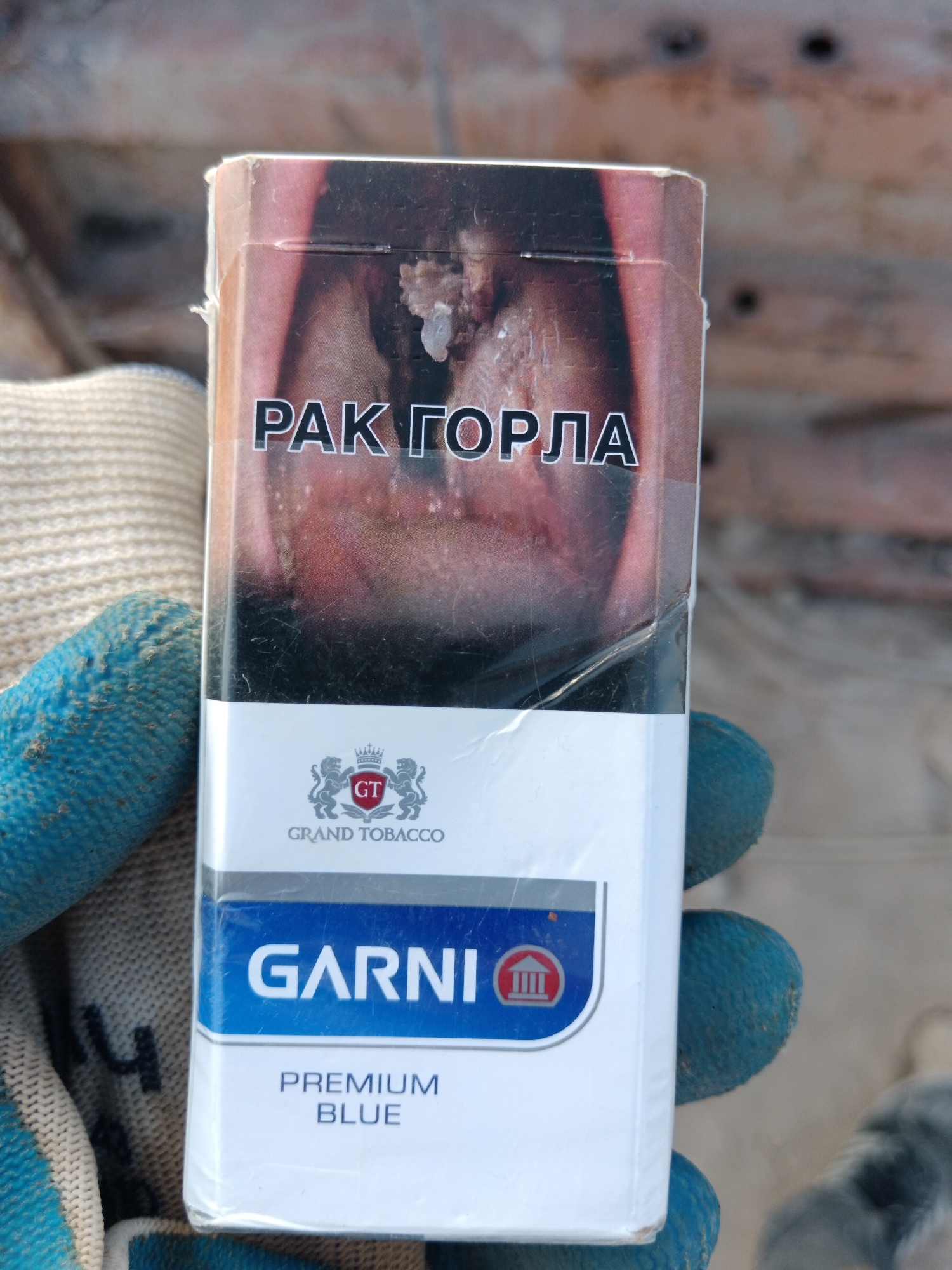 The image shows a close-up of a pack of "GARNI PREMIUM BLUE" cigarettes. The pack has a warning label on the top featuring a graphic of a diseased throat with the text "РАК ГОРЛА" (Throat Cancer) in Russian. Below this, the "GT GRAND TOBACCO" logo is visible, followed by the brand name "GARNI" and the product description "PREMIUM BLUE". The cigarette pack is held in a gloved hand, with a portion of a beige knitted glove and a blue textured glove visible on the left. The background appears to be a rough, textured surface, possibly wood or concrete, with some dark circular marks. The scene is set outdoors in Aragatsavan, Armenia, where the pack of cigarettes is being held.