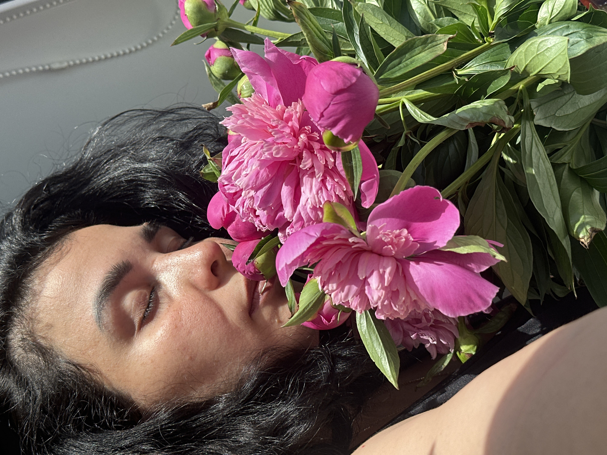 The image captures a close-up scene in the city of Unknown, Unknown, featuring an adult female with her eyes closed, partially obscured by a vibrant bouquet of peonies. Her dark, wavy hair is visible on the left and top of the frame. Her face, in profile, exhibits an olive skin tone with visible pores and texture, and is gently nestled among the flowers, suggesting she is smelling them. The bouquet consists of multiple fuchsia-pink peonies, some fully bloomed with ruffled petals and others as tight buds, accompanied by numerous healthy green leaves and stems. A strong, directional light source, likely natural sunlight, illuminates the subject from the top right, creating bright highlights on her skin, hair, and the flowers, while casting a distinct shadow across her left shoulder and arm, where a dark garment is visible. The background on the right is a light-toned, out-of-focus surface, revealing a vertical element with small, evenly spaced, bead-like details, consistent with a window blind or similar structure.