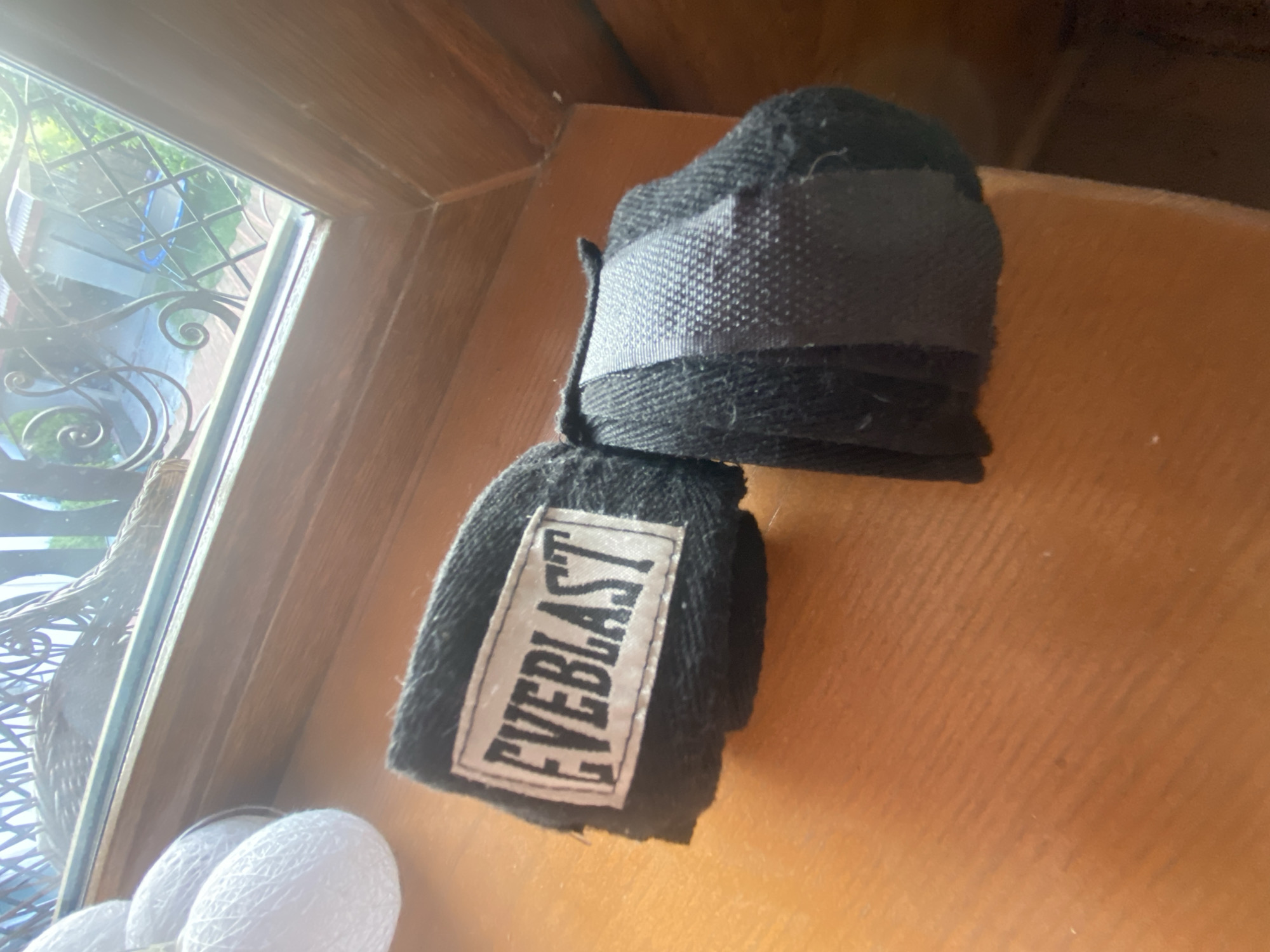 Two black fabric hand wraps are positioned on a light brown wooden surface, likely a windowsill, under bright daylight. The wrap on the left displays a white rectangular label with the black text "EVEBLAST" prominently displayed. The wrap on the right is also rolled, revealing a gray fabric strap or velcro closure. To the far left, several white spherical objects made of tangled string are partially visible. Beyond the wooden surface, a window reveals an outdoor scene with an ornate dark metal railing, green foliage, a partial view of a light-colored building with a roof, and a distant blue circular object, possibly a trampoline. The location is in the city of Unknown, Unknown.