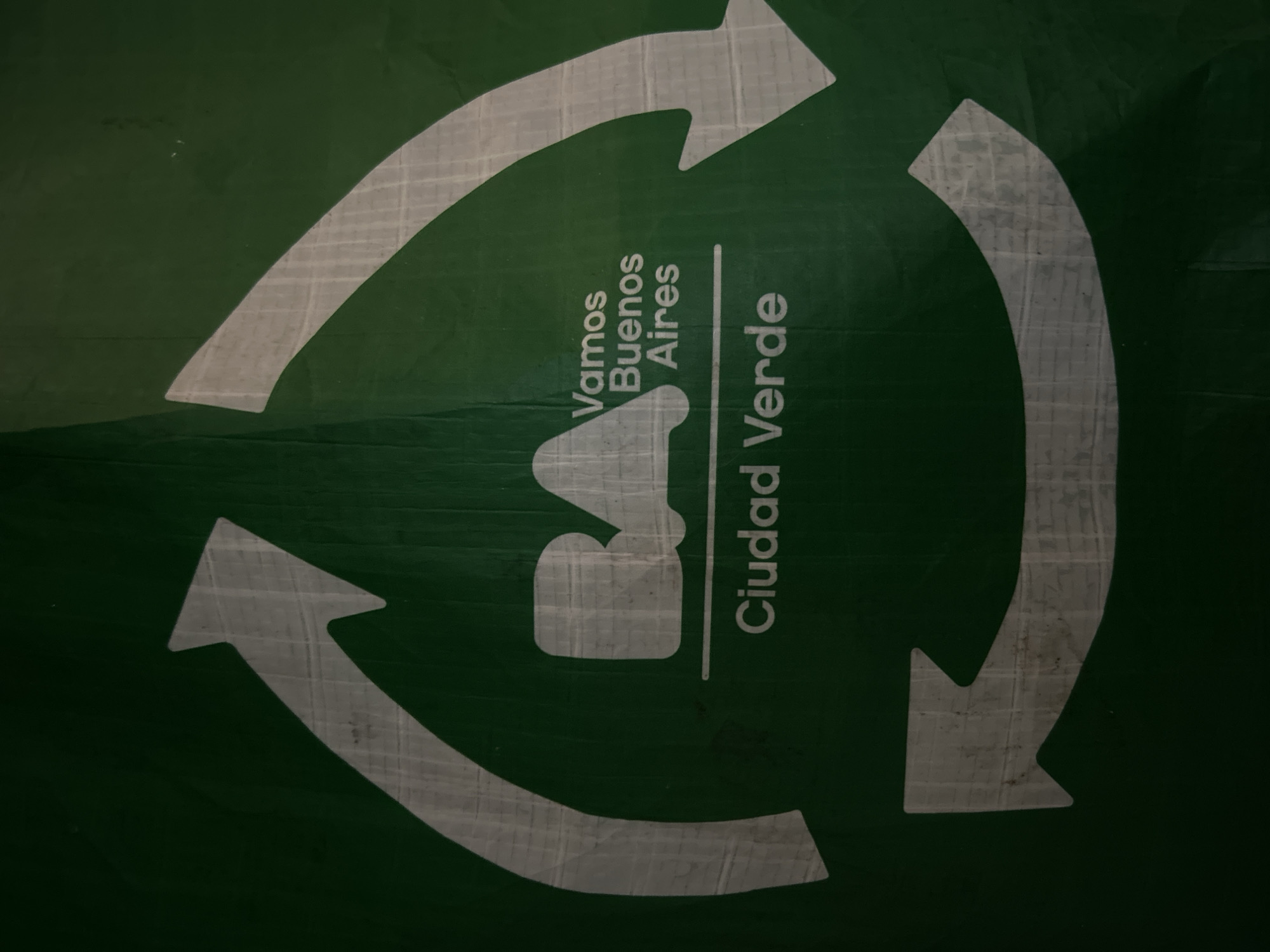 This is a close-up, eye-level photograph of a green banner featuring a white recycling symbol with text. The banner displays the stylized logo "BA" with the words "Vamos Buenos Aires" above and "Ciudad Verde" below, all in white text. The recycling symbol is composed of three curved arrows forming a circular shape. The banner appears to be made of a flexible material and has a slightly creased texture. The lighting suggests it is outdoors or in a well-lit indoor space. The image does not depict any specific identifiable landmarks of Buenos Aires, Argentina.