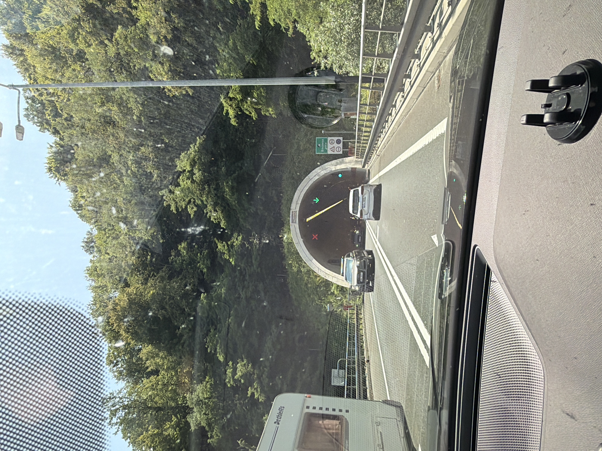 The image is taken from inside a vehicle approaching a tunnel entrance on a sunny day. The foreground shows the dashboard of the vehicle with a phone mount. A white caravan with "Dethleffs" branding is visible to the left. In front of the vehicle, a grey Audi SUV is driving in the right lane, and a white car is in the left lane, both heading towards the tunnel. The tunnel entrance is illuminated, and traffic lights and signage are visible. The tunnel is surrounded by a lush green, tree-covered hillside. A street lamp stands on the right side of the road. A green directional sign indicates "Brezovica 200 m". The road has two lanes in each direction, separated by white lines, and a metal guardrail runs along the right side. The sky is clear and blue. The location is identified as Durmanec, Croatia.