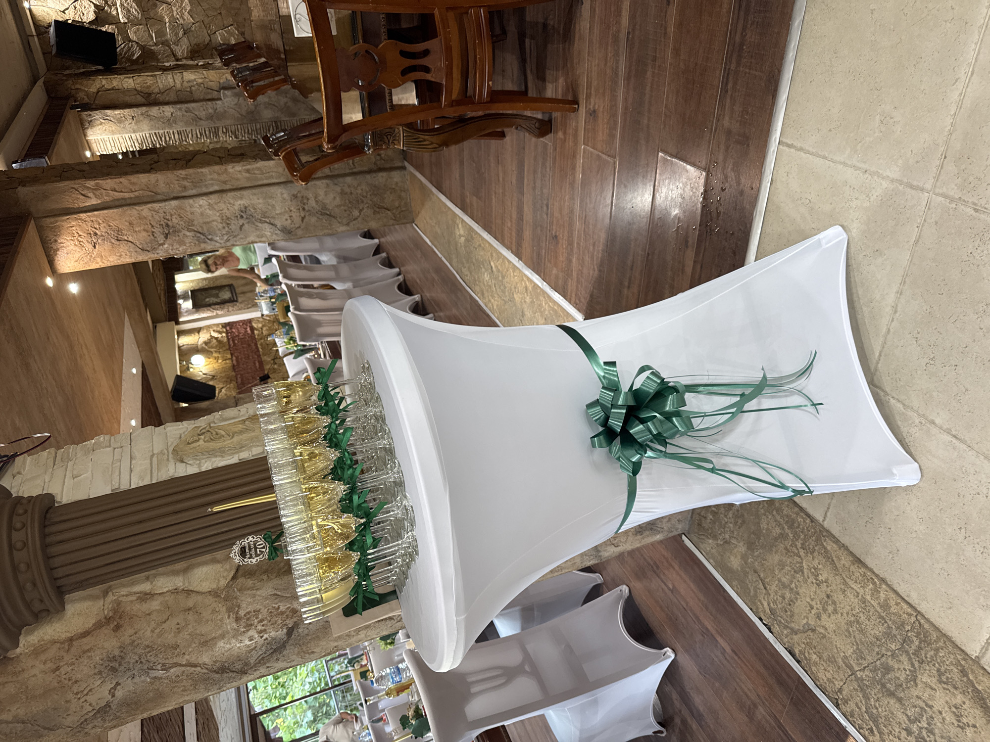 The image depicts an indoor scene, likely a venue for a celebration, possibly in Sredets, Bulgaria. In the foreground, a tall, white-covered table displays a row of champagne flutes filled with a golden liquid. A decorative green ribbon bow is tied around the base of the table. Behind this table, to the left, another similar table is set with white chair covers. In the mid-ground, a woman in a light green top is visible in the background, near tables and chairs. The walls are decorated with stone and wood finishes, and there are wooden pillars. Further back, dining tables and chairs are arranged. The floor is a combination of dark wood and light-colored tile. The overall impression is of a prepared event space.