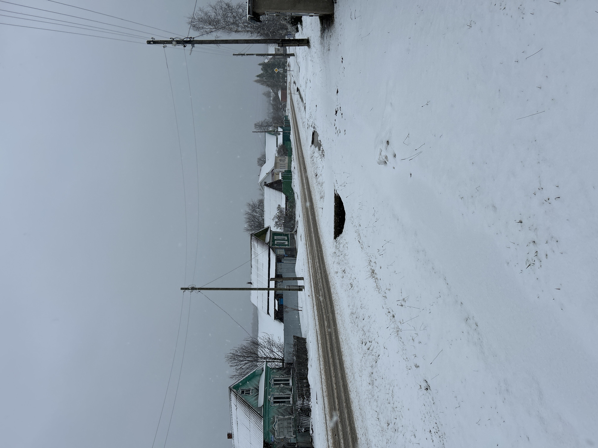 The image depicts a snow-covered street in Уральское, Russia. The street is flanked by houses, some of which appear to be older wooden structures. Utility poles line the side of the road, with power lines visible against the overcast sky. The road itself shows evidence of tire tracks, suggesting recent vehicular traffic through the snow. The environment is characterized by a winter landscape with a uniform layer of snow covering most surfaces. There are no people visible in the frame.