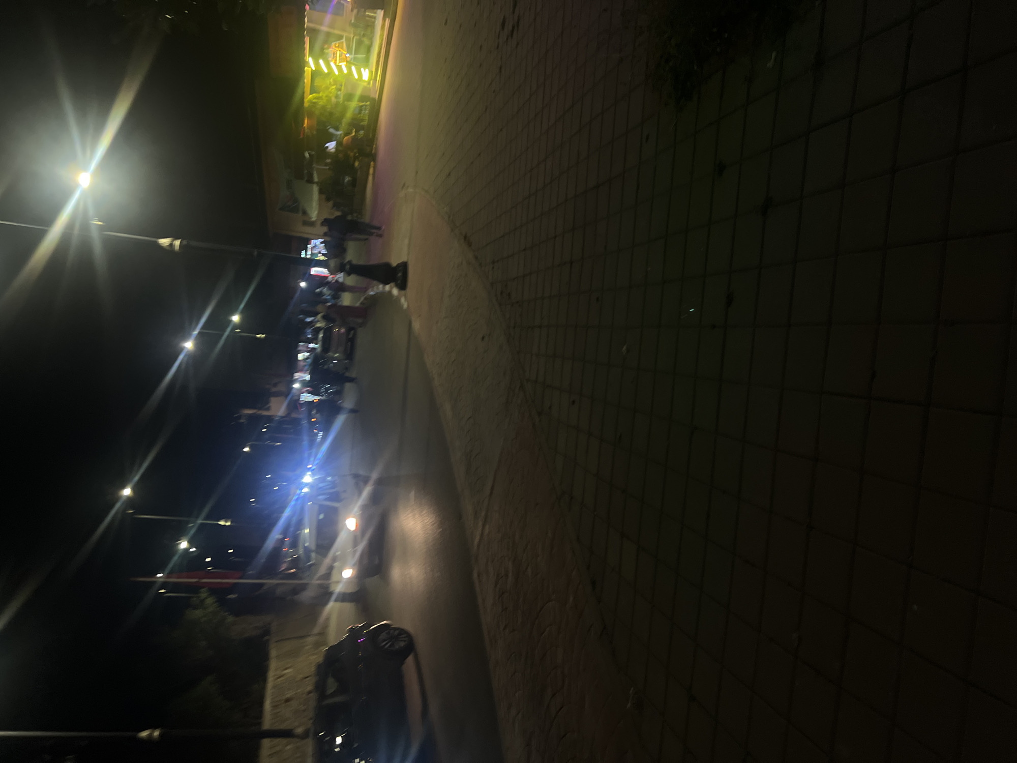 The image shows a street scene in Bin El Ouidane, Morocco at night. Street lights illuminate a row of parked scooters and motorcycles along a curved sidewalk. A group of people is gathered near a brightly lit building or shop. The walkway is paved with interlocking blocks.