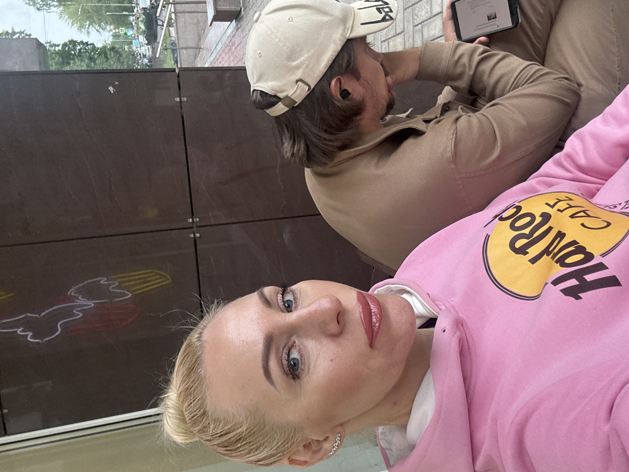 A blonde woman with blue eyes and pink lipstick is in the foreground of the image, looking towards the camera. She is wearing a pink hoodie with a "Hard Rock Cafe" logo. To her right, a person wearing a tan hoodie and a tan baseball cap is seated, looking down and holding a smartphone. The background appears to be an urban setting, with a brown tiled wall, some greenery, and a street with vehicles visible through a glass pane. The lighting suggests it is daytime.