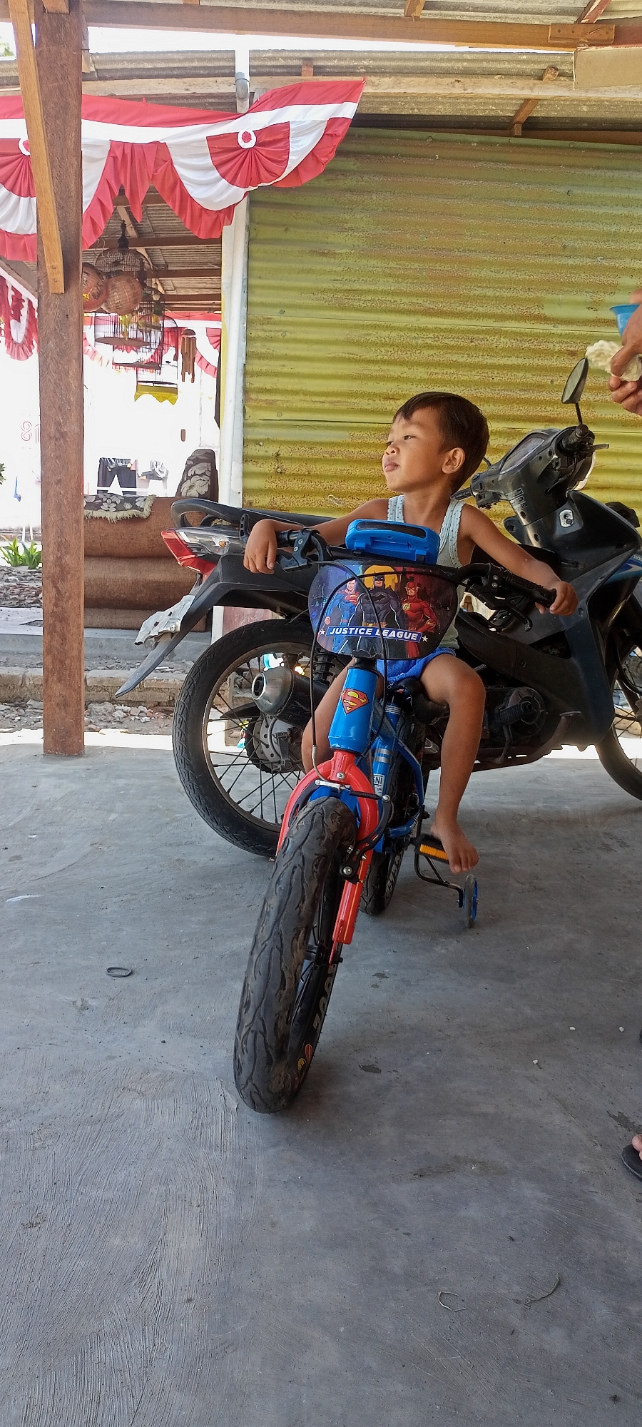 A young boy is sitting on a motorcycle toy. The boy is wearing a white tank top and blue shorts. The motorcycle toy is blue and red with images of Justice League characters on the front. The boy's legs are extended, and his feet are resting on the ground. The motorcycle toy has training wheels. In the background, there is a corrugated metal wall painted yellow and a wooden structure with red and white bunting hanging from it. There are also several bird cages visible. The scene appears to be outdoors, possibly in a residential area or a market. The ground is concrete.