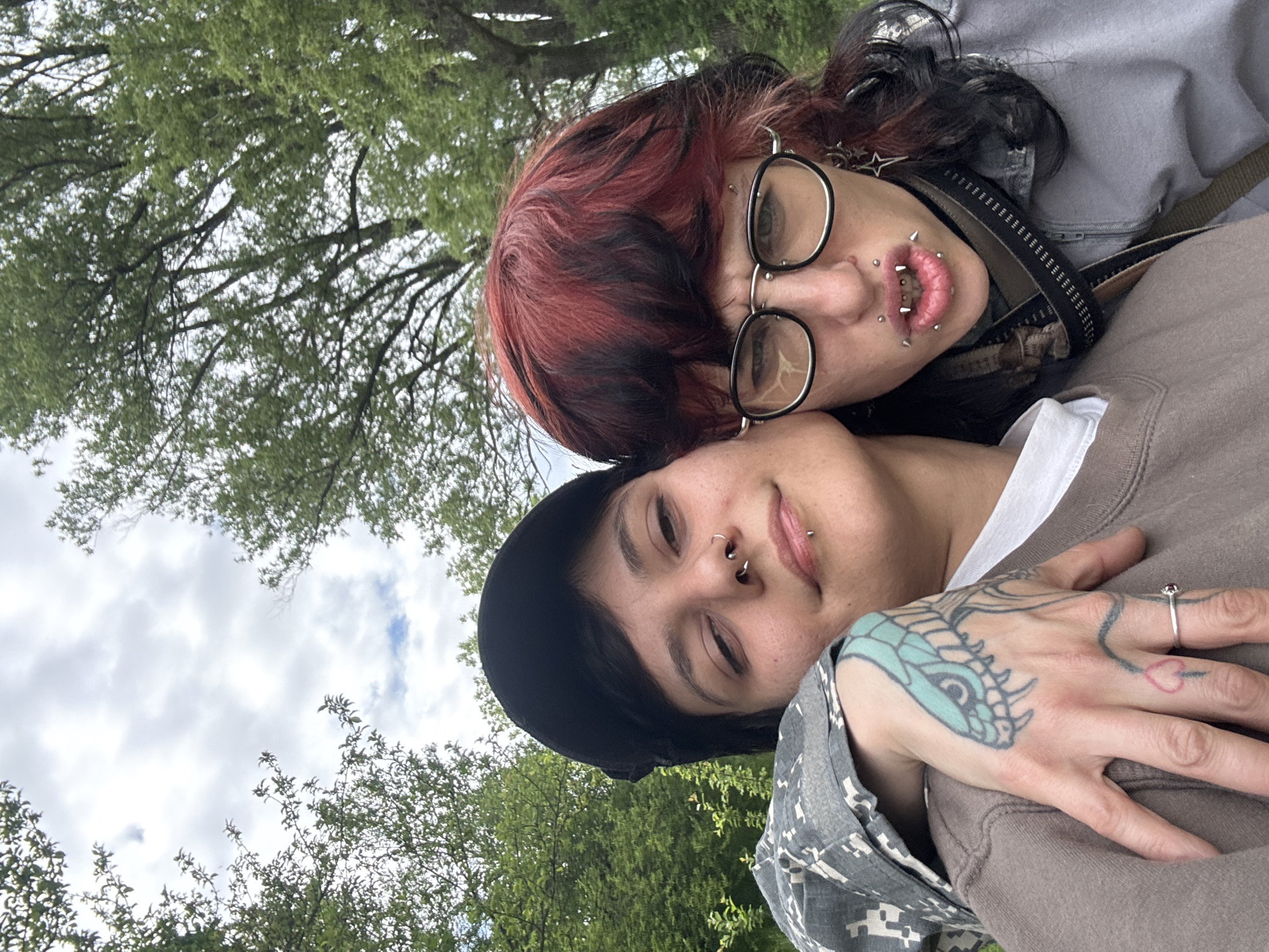 Two individuals are visible in the image, likely a selfie or taken at close range. The top individual has red and black hair, wears glasses, and has multiple piercings on their face, particularly around their lips. They are wearing a grey jacket with a strap visible across their body. The individual beneath them has a black hat, a nose piercing, and a tattoo on their left hand. They are wearing a grey and white camouflage patterned jacket underneath a brown sweater. The background consists of green trees and a cloudy sky. The location cannot be determined without additional information.
