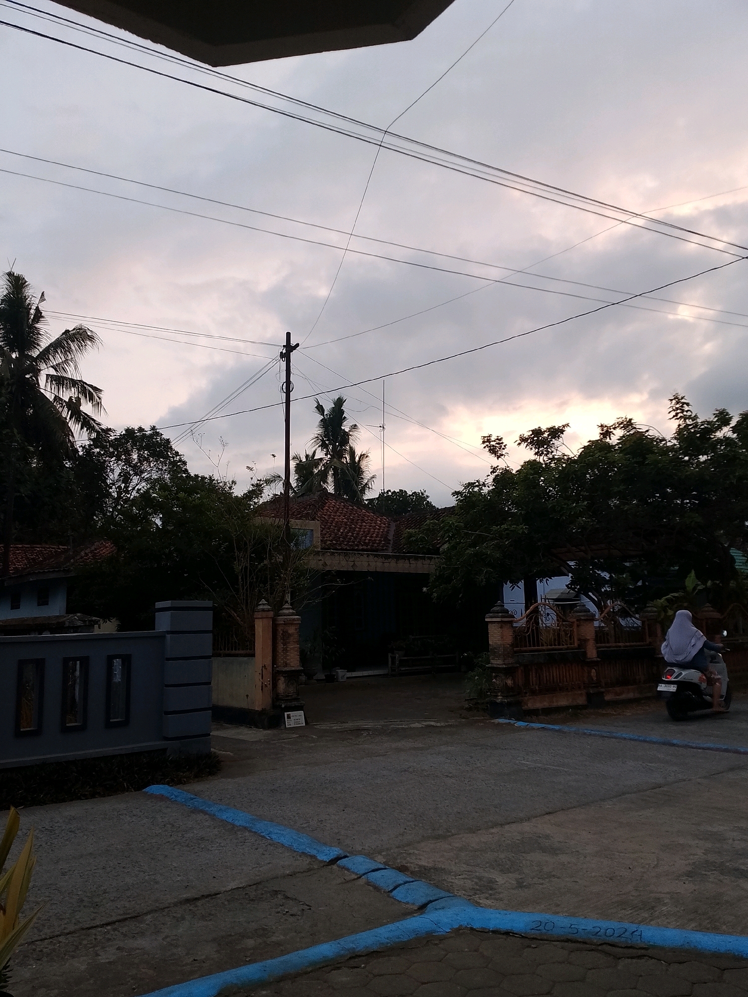 The image captures a street scene in Gebang, Indonesia, likely during the late afternoon or early evening, given the soft, diffused light. A person wearing a hijab is riding a scooter away from the camera towards the right side of the frame. The scooter is light gray and has a license plate that appears to read "PA 3030 RY". The scooter is on a paved road, which is marked with blue painted lines.
On the left side of the road, there is a low, modern-style gray wall with three rectangular openings, possibly decorative or for ventilation. Behind this wall, palm trees and other lush green trees are visible. Further back, a house with a red tiled roof is partially obscured by the foliage.
To the right of the house, there's a brown fence with intricate metalwork. Beyond the fence, more trees and another house with a red tiled roof are seen. The sky above is overcast with soft, muted colors, suggesting either dusk or dawn. Several power lines crisscross the sky, and a utility pole stands prominently in the mid-ground. The overall impression is of a residential area in a tropical or subtropical climate.