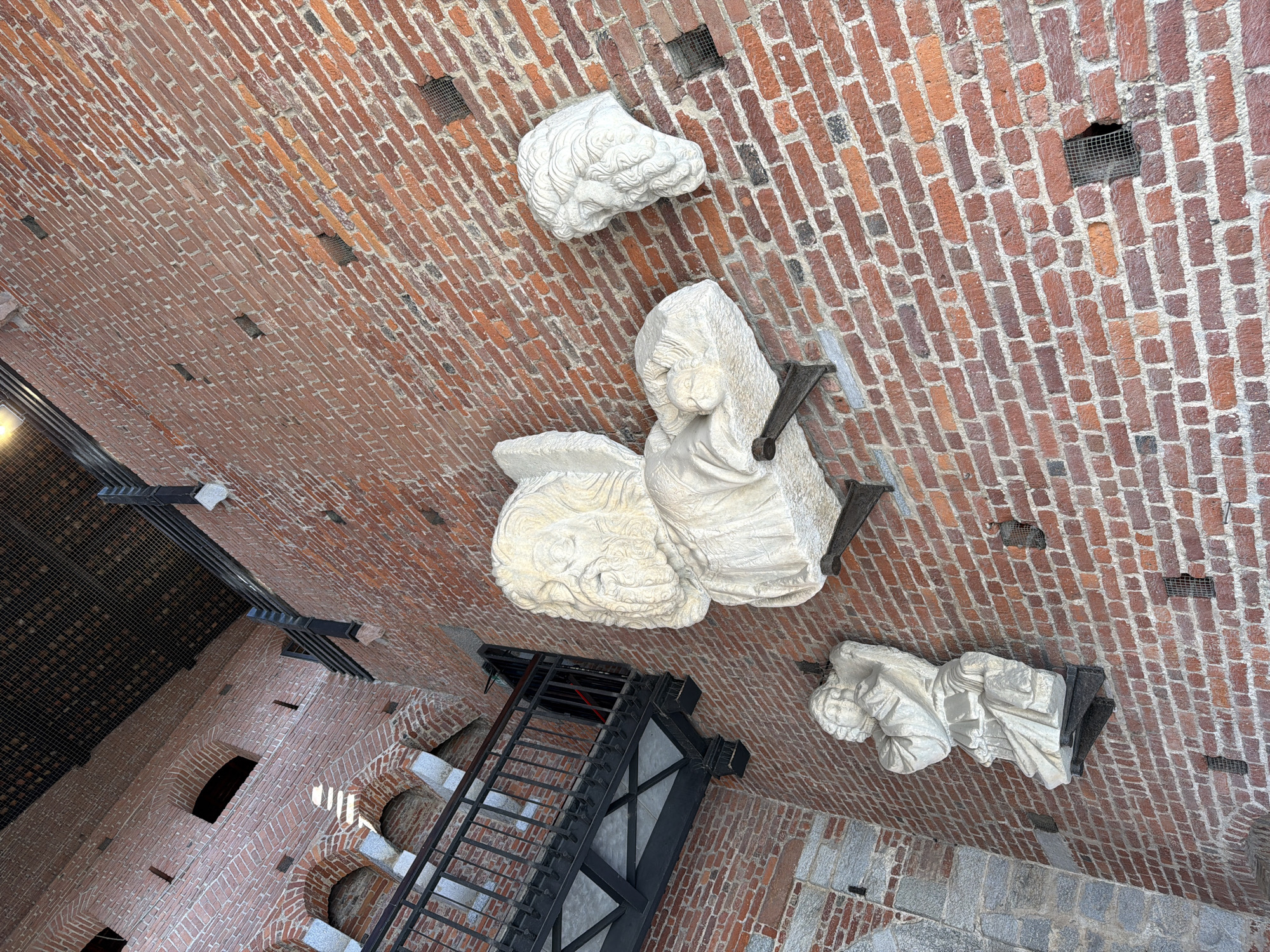 The image presents an upward view of a high, vaulted ceiling and adjacent structures, likely within a historic building in Milan, Italy. The primary subjects are three white stone or marble sculptures affixed to the red-brick ceiling.
One sculpture, located in the upper-middle, depicts a head or bust with finely detailed, wavy hair or a mane. Below and slightly to the left, a larger sculpture portrays a draped figure, possibly reclining or seated, holding what appears to be an infant or small child. This piece is supported by a dark metal bracket. A third, similarly draped figure in a recumbent pose is visible towards the lower right, also supported by dark metal elements.
The ceiling itself is composed of reddish-brown bricks laid in a diagonal pattern, with visible mortar lines. Several small, square openings covered with dark metal mesh are interspersed across the brickwork. Towards the lower left, a dark metal staircase or viewing platform with a grid-like floor descends into the space. Further down, arched openings or windows are visible in the brick wall, with rays of sunlight streaming through one of them, indicating a bright, clear day. The architectural style, combined with the presence of historic sculptures and contemporary access structures, suggests a significant cultural or religious edifice. No text is visible in the image.