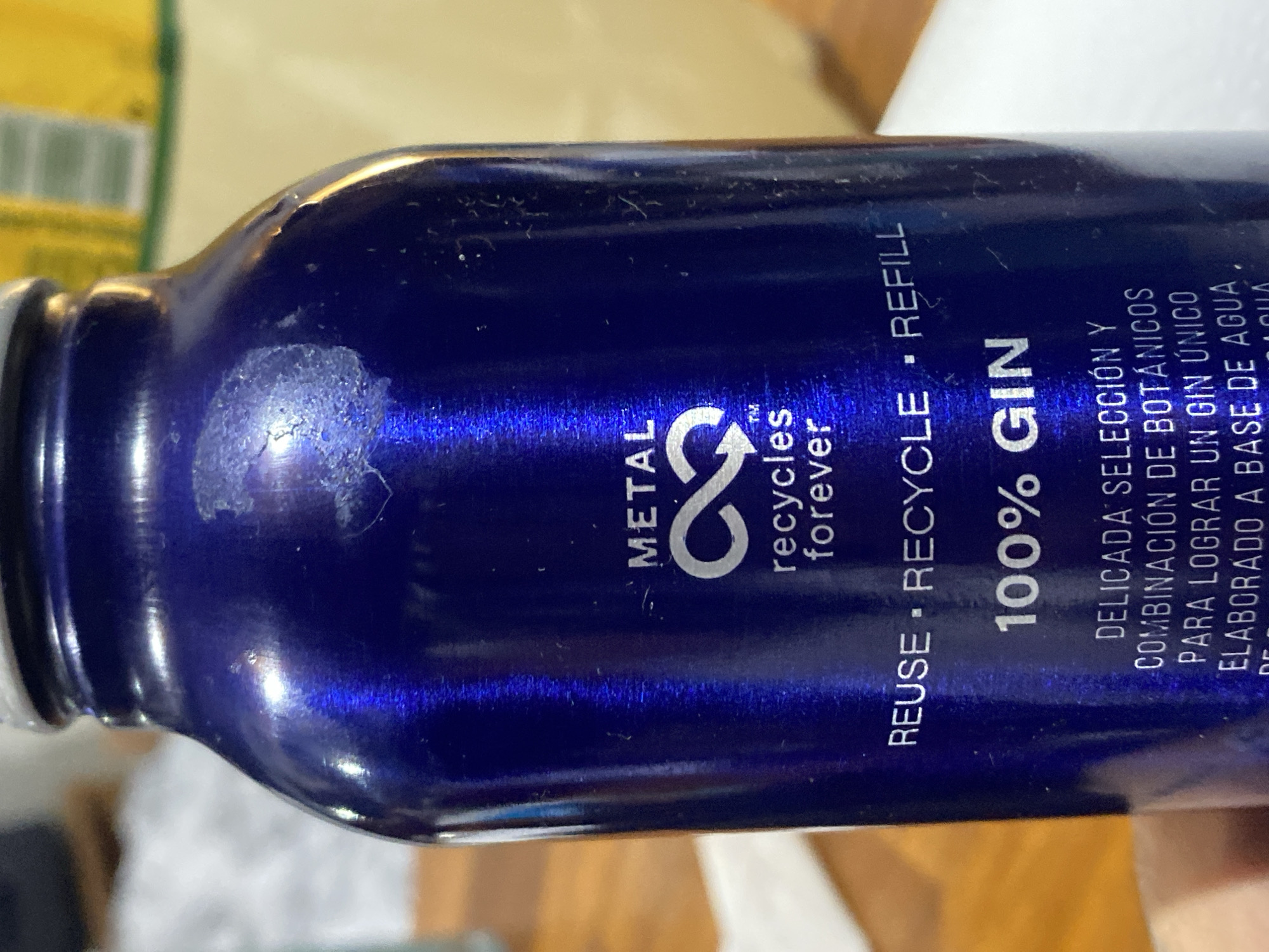 An eye-level, close-up shot captures a blue metallic bottle, likely made of aluminum, lying horizontally on what appears to be a light-colored surface, possibly a wooden table or countertop. The bottle is a deep, iridescent blue with a textured finish, showing some visible scuffs, scratches, and a faded, circular mark near the neck, suggesting prior use or handling.
The bottle's design is prominent with white text and a silver logo. A recycling symbol, stylized as two arrows forming an '8', is visible, accompanied by the text "METAL" above it and "™ recycles forever" below. Further along the bottle, the words "REUSE • RECYCLE • REFILL" are printed vertically. The contents are identified as "100% GIN".
Below the gin identification, there is Spanish text that reads: "DELICADA SELECCIÓN Y COMBINACIÓN DE BOTÁNICOS PARA LOGRAR UN GIN ÚNICO ELABORADO A BASE DE AGUA". This translates to "DELICATE SELECTION AND COMBINATION OF BOTANICALS TO ACHIEVE A UNIQUE GIN MADE WITH WATER BASE". The use of Spanish aligns with the given location context of Buenos Aires, Argentina.
The background is out of focus, revealing hints of yellow and other muted tones, suggesting an indoor setting. The overall impression is that of a product shot highlighting the reusable and recyclable nature of the bottle and its gin contents.