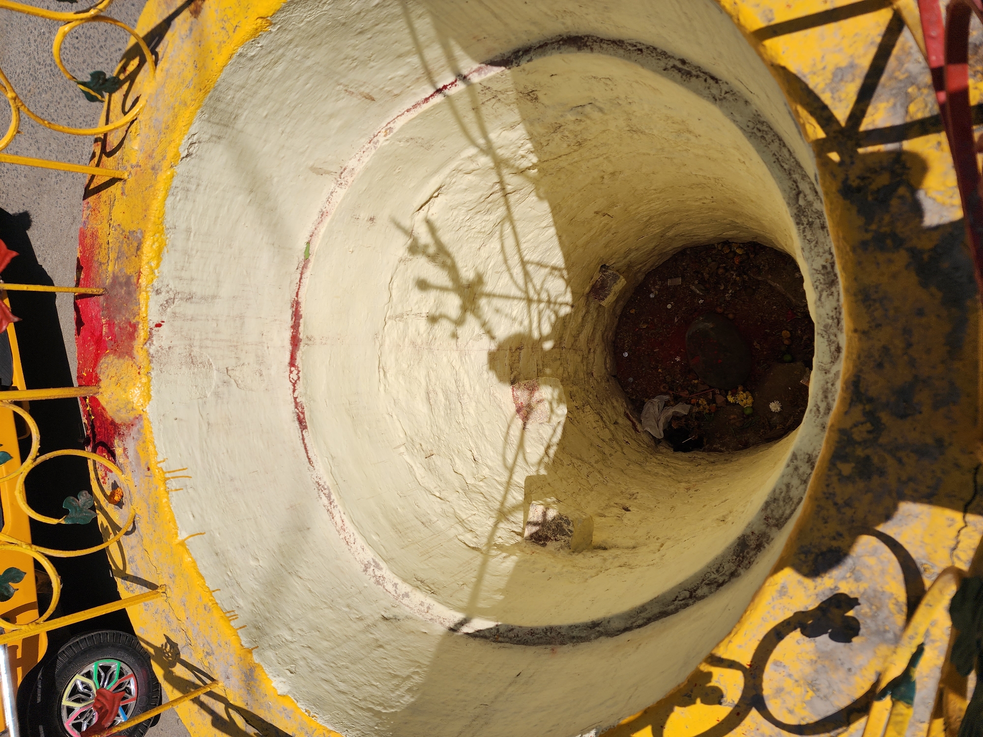 The image presents a top-down view into a deep, cylindrical, well-like structure in an outdoor setting. The location in the city of Unknown, Unknown, is not discernible.
The structure's exterior rim is painted yellow, exhibiting wear, chipping, and several distinct drips of red paint. A yellow metal railing with decorative scrollwork and small green leaf accents partially encircles the well's opening. In the upper left corner, the top of a yellow vehicle, likely an auto-rickshaw, is partially visible, showing a wheel with a multi-colored hubcap. The ground immediately adjacent to the well and vehicle appears to be grey asphalt or concrete.
The interior of the well is painted a textured, cream-to-pale-yellow color, with irregular reddish-brown horizontal bands visible on its rough surface. The well narrows towards its bottom, where a dark, earthen base is visible. Within this base, various items are scattered, including small white and yellow flowers, numerous coins, and a piece of white plastic or fabric. A dark, possibly conical object rests near the center of the base.
Strong sunlight casts prominent shadows. Shadows from the metal railing are visible on the exterior rim and the upper interior of the well. A human shadow, consistent with a person holding a camera or phone, is cast onto the well's interior wall from the upper left. Adjacent to this, a distinct, multi-pronged shadow, resembling a ceremonial lamp or decorative stand, is also cast onto the well's interior. No individuals are directly visible. The scene suggests a site of ritual or offering.