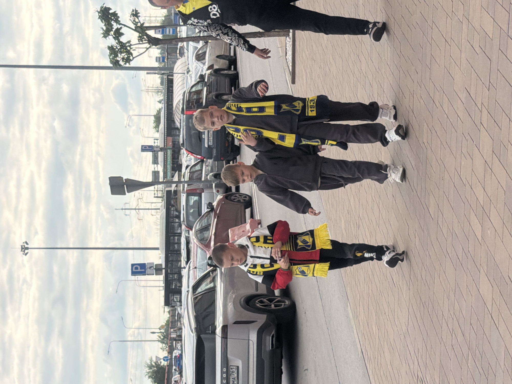 Three boys and a man stand on a brick-paved sidewalk next to a parking lot full of cars. The boys wear scarves with yellow fringe and the word "OB" or "JOEB" and "1930" printed on them. The man wears black athletic pants and a black jacket with yellow and white trim. A sign with a "P" inside a blue square indicates parking. Car license plates are visible, including one that reads "COO MA 761". The scene is in Rostov-on-Don, Russia.