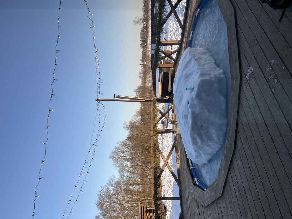 The image depicts an outdoor recreational area in Shipulino, Russia, under clear, sunny winter conditions. The foreground features a weathered wooden deck with an octagonal-shaped, in-ground pool. This pool is largely occupied by a substantial, irregular mass of white snow and ice, with a small, light green plastic bottle partially embedded in its surface. Scattered pieces of ice are visible on the wooden deck near the pool.
A rustic wooden fence, featuring crisscross bracing and adorned with a continuous evergreen garland, separates the deck area from a snow-covered landscape. Beyond the fence, the ground is uniformly covered in white snow. Visible in this midground are a dark, round wooden tub with integrated benches, and a tall wooden pole with a metallic chimney pipe affixed to its side. A small wooden structure with horizontal slats is partially visible to the left.
The background consists of a dense line of bare deciduous trees, followed by an open, snow-covered field. In the far distance, a cluster of buildings from a settlement is discernible, including a prominent structure with a light-colored steeple or tower. The sky above is clear and bright blue, spanned by two parallel strings of decorative incandescent fairy lights. No individuals are present in the scene.