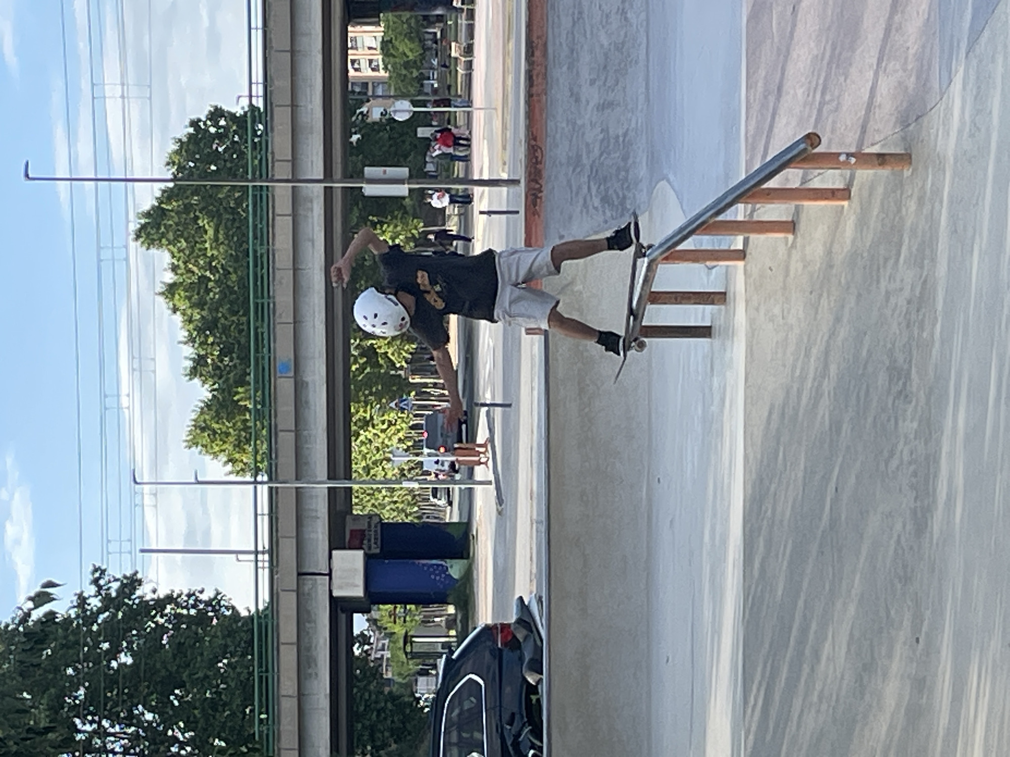 This image depicts a skateboarder performing a trick on a metal grind rail in an outdoor skate park. The scene is set on a bright, sunny day, likely in the late morning or early afternoon, under a clear blue sky with a few wispy clouds.
The main subject is a skateboarder, wearing a white helmet, a dark t-shirt with a light-colored graphic design, light-colored shorts, and dark shoes. They are in mid-action, with their skateboard balanced on an inclined metal rail that is supported by wooden steps. The skateboarder’s body is angled, and one arm is extended for balance, conveying focus and control during the trick.
The background reveals an urban or suburban environment. A line of lush green trees borders the skate park, behind which an elevated road or bridge structure with its supporting pillars and overhead power lines is visible. Distant buildings can be seen under the bright sky. On the ground level, a dark-colored car is partially visible, parked near a blue public trash or recycling bin. Faint text on the bin, possibly in Catalan, includes phrases like "PURA I SIMPLE" and "RECYCLE." The skate park itself features smooth concrete surfaces and hints of other ramps or bowls. The overall impression is one of active recreation in a sunny public space in Santa Perpètua de Mogoda, Spain.