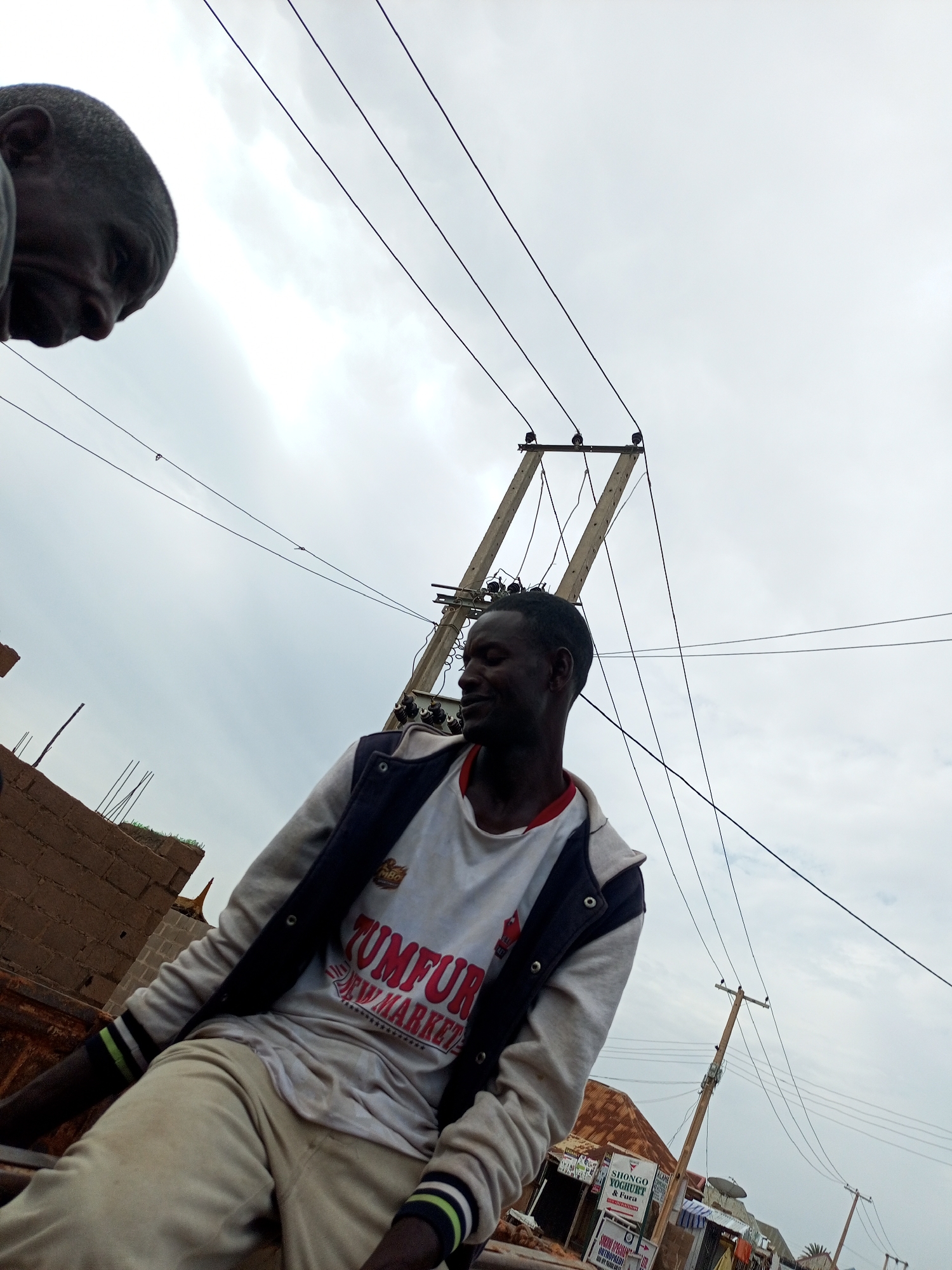 A low-angle outdoor shot shows two Black men against a cloudy sky, with utility poles and wires prominently featured. The man on the left, partially visible in the top left corner, is looking down. The man on the right, in the foreground and center, is looking to his left with his eyes closed. He is wearing a white t-shirt with red lettering that reads "TUMFUR NEW MARKET" and a grey and dark blue jacket. He is also wearing light-colored pants. To the left of the man on the right, a brick wall under construction is visible, with rebar protruding from the top. In the background, several utility poles with wires are present, and a low-rise building with a corrugated metal roof can be seen. A sign for "SHONGO YOGURT & FURA" is also visible on the building. The overall impression is of an urban or semi-urban environment with ongoing construction.