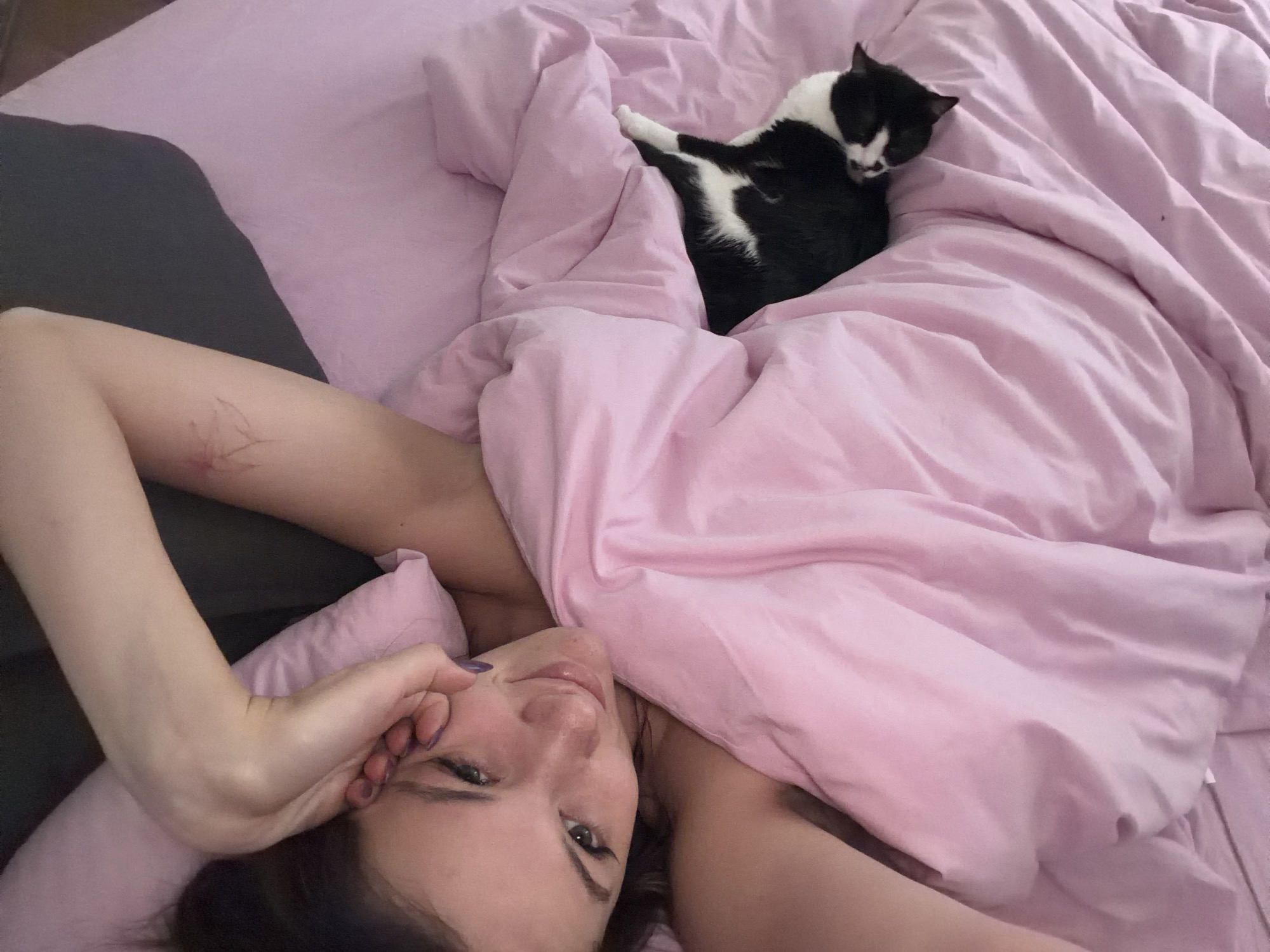A woman lies in a bed with pink bedding and a black and white cat. The woman's face is visible, and she has dark hair and light skin. Her left arm is raised near her face. The cat is nestled under the bedding to the right of the woman. A grey pillow is to the left of the woman. The location is not evident in the image.