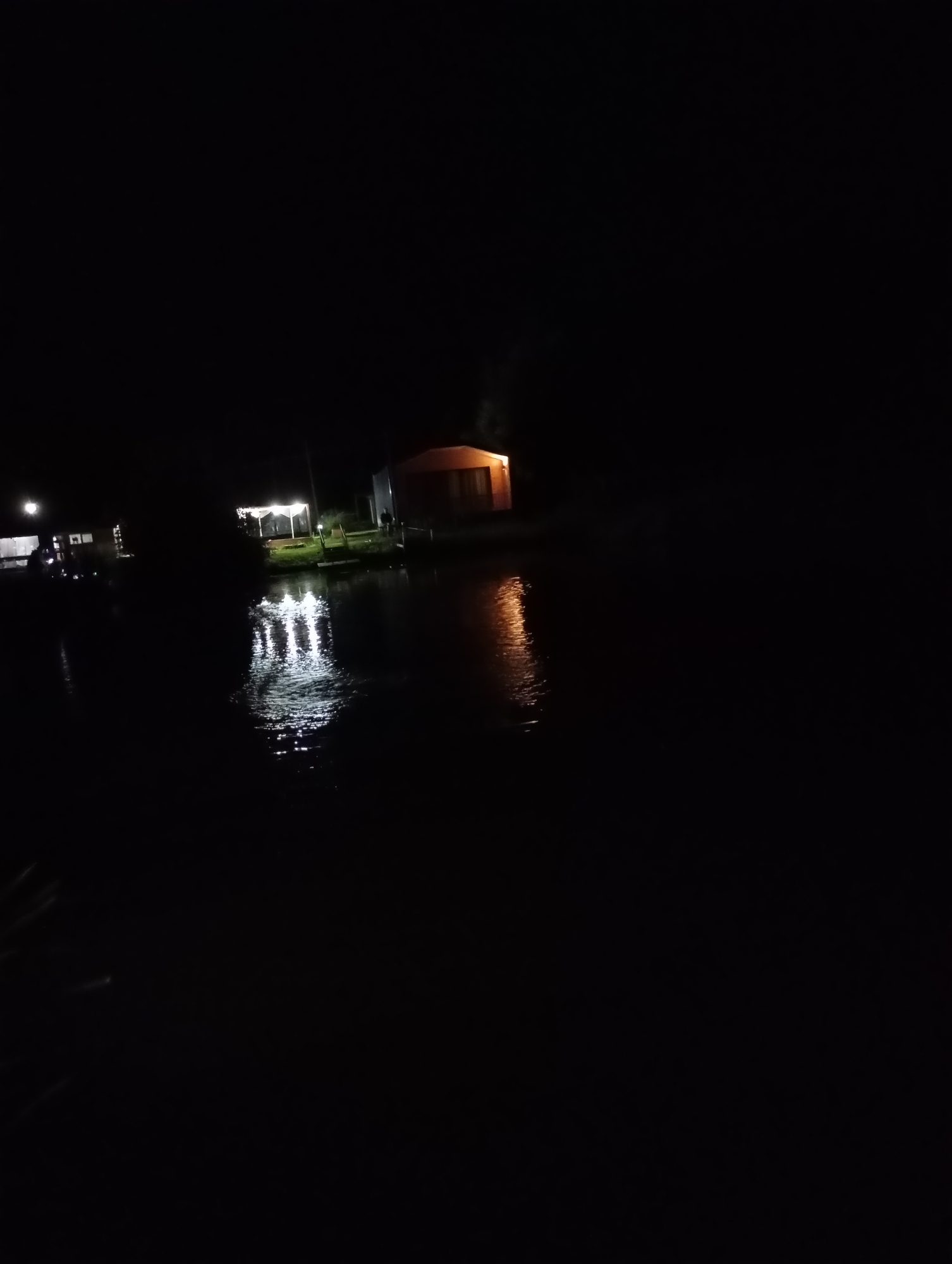 A nighttime scene shows a body of water reflecting artificial lights from structures on the shore. On the left, a building with lights is partially visible. In the center, a covered outdoor seating area with bright lights is illuminated, with reflections on the water. To the right, a single-story building with orange-toned lights is visible, its reflection also present on the water. The overall environment is dark, with the water occupying the foreground and the structures situated along the shoreline. The location is identified as Malyi Bukor, Russia.