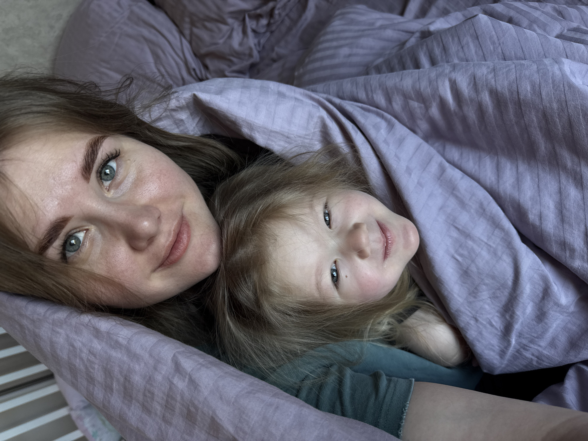A selfie captures a woman and a young child lying in a bed with lavender-colored bedding. The child is nestled under a duvet. Both are looking at the camera and smiling. The location is not discernible from the image; the city of Ферма, Russia, cannot be confirmed as the location.