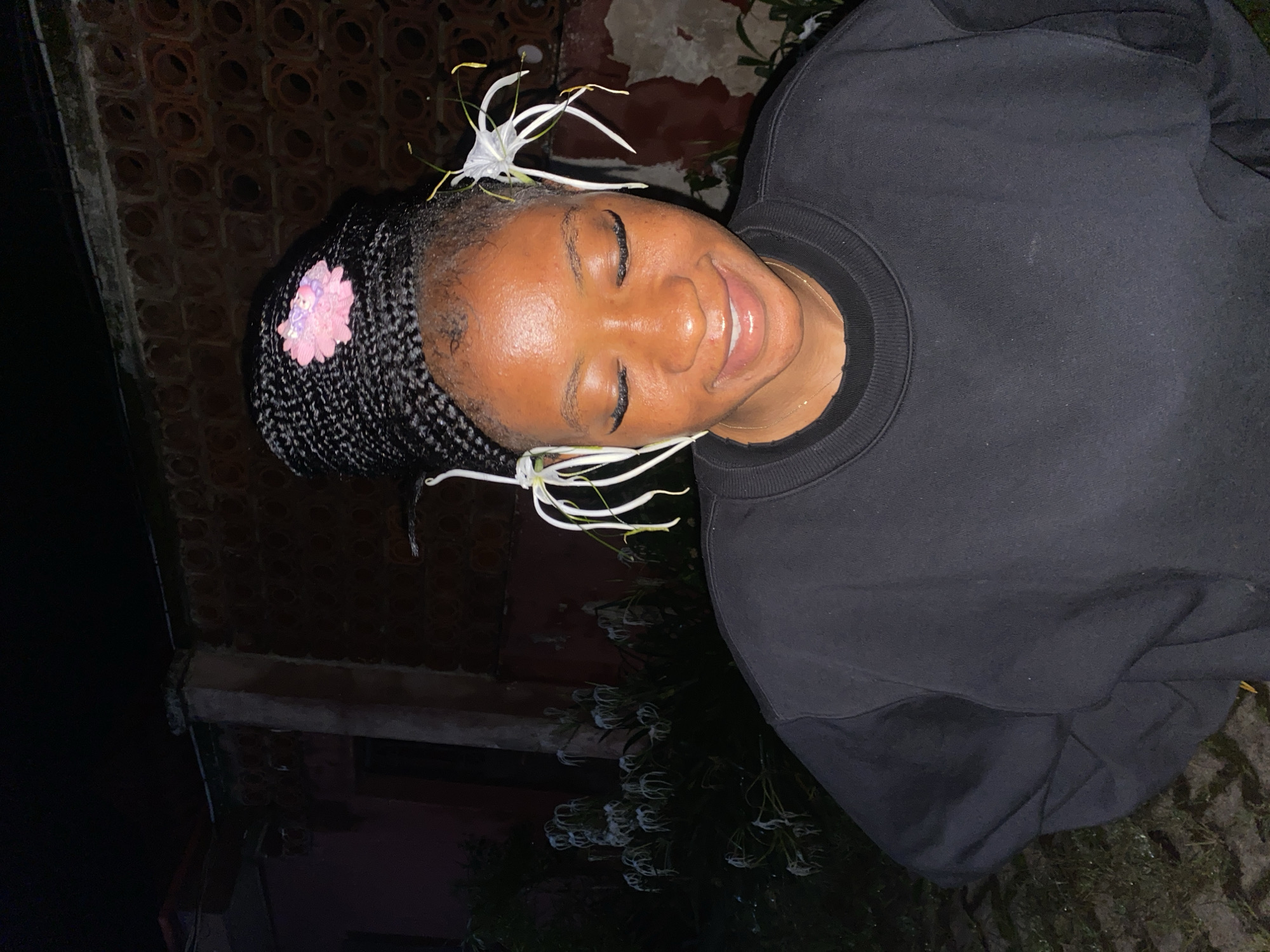An undated, full-body shot captures a young Black woman with her eyes closed and a slight smile, adorned with two white spider lilies placed near her temples and a pink flower clip on top of her braided hair. She is wearing a black crewneck sweatshirt and a thin gold necklace. The background suggests an outdoor setting at night, with a brick wall and lush greenery visible. The context of Jos, Nigeria, is not evident from the image.
