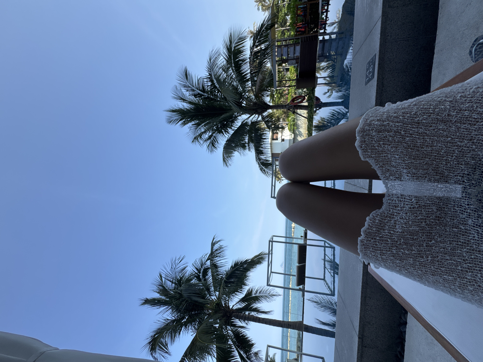 Here is a forensic description of the image:
The image shows a view of the sky and palm trees, as seen from a low vantage point. A pair of legs, presumably belonging to a person, are visible in the foreground. The legs are pale and smooth, and they are crossed at the knees. The person is wearing a white, crocheted skirt or dress. The scene appears to be near a body of water, likely a pool or the ocean, based on the color and reflections visible in the background. The sky is clear and blue. The palm trees are tall and have green fronds. The overall impression is of a relaxing, tropical setting. The location is Unknown, Unknown.