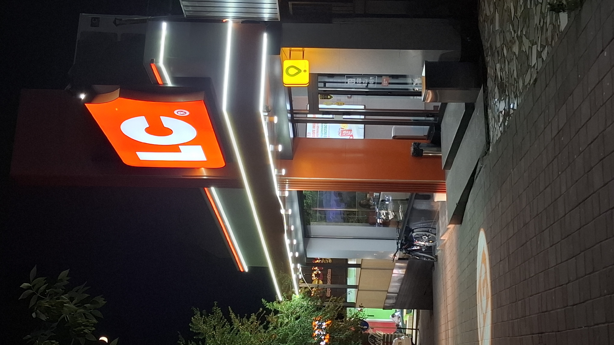 The image shows the entrance of an LC Waikiki store at night in Shymkent, Kazakhstan. The store has a prominent orange sign with the LC logo in white. White LED lights illuminate the storefront, accentuating its modern design. To the left of the entrance, a tree with green leaves is visible, and a crescent moon is in the dark night sky. In front of the store, a bicycle is parked on a paved walkway. The walkway is illuminated by a circular projection of the LC logo. A bright yellow sign with a location pin icon is mounted above the entrance. Inside the store, a person can be seen behind a counter. The overall scene depicts a well-lit commercial establishment in an urban setting at night.