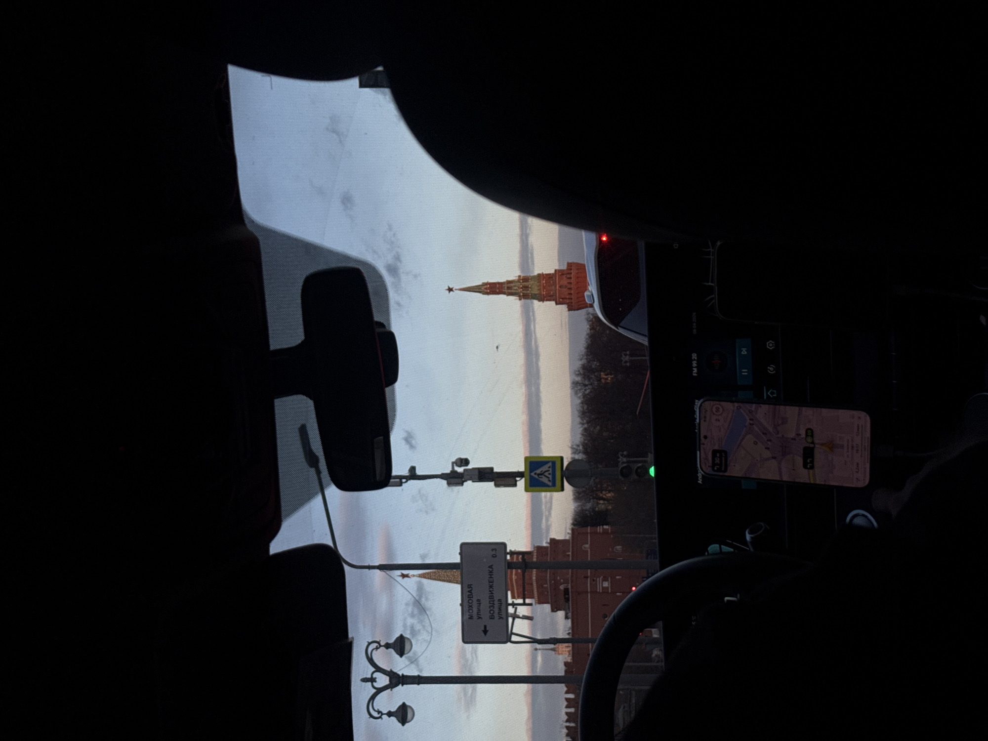 The photo, taken from inside a car, offers a view of the Moscow Kremlin, including the iconic spire topped with a red star. The car's interior, including the rearview mirror, dashboard, and steering wheel, is visible, framing the external scene. Outside, street signs indicate "МОХОВАЯ" (Mokhovaya) street and "ВОЗДВИЖЕНКА 0.3" (Vozdvizhenka 0.3) street, pointing towards the direction of the Kremlin. A pedestrian crossing sign is also visible. A smartphone displaying a map, presumably for navigation, is mounted on the dashboard. The street lamps add detail to the urban landscape. The time on the phone is 7:30 pm.