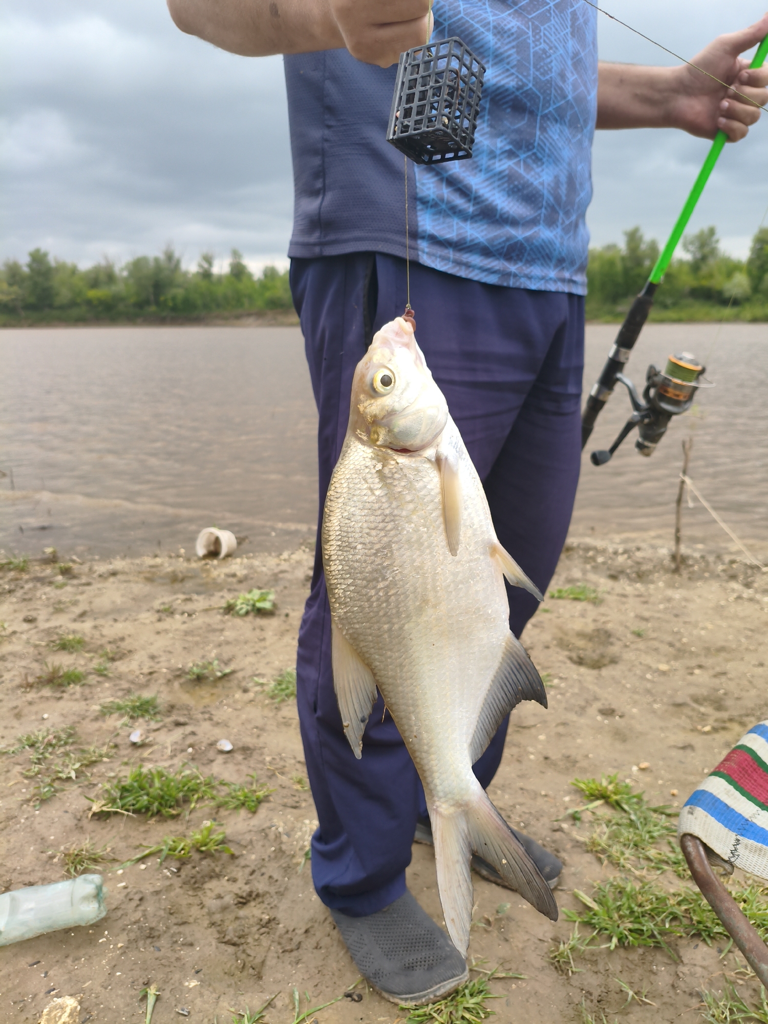 A person, dressed in a dark blue t-shirt with a geometric blue pattern and dark blue trousers, stands on a muddy bank next to a body of water. The person is holding up a freshly caught fish by a fishing line, which is attached to a black, square feeder cage. A green fishing rod with a reel is visible in the person's right hand, resting against their shoulder. The background shows a treeline and a cloudy sky. On the ground near the person are some scattered items including a crushed plastic bottle and some green grass. A portion of a striped chair seat is visible in the lower right corner of the frame. The scene appears to be a casual fishing outing, possibly in the rural area of Ақсуат ауылдық округі, Kazakhstan.