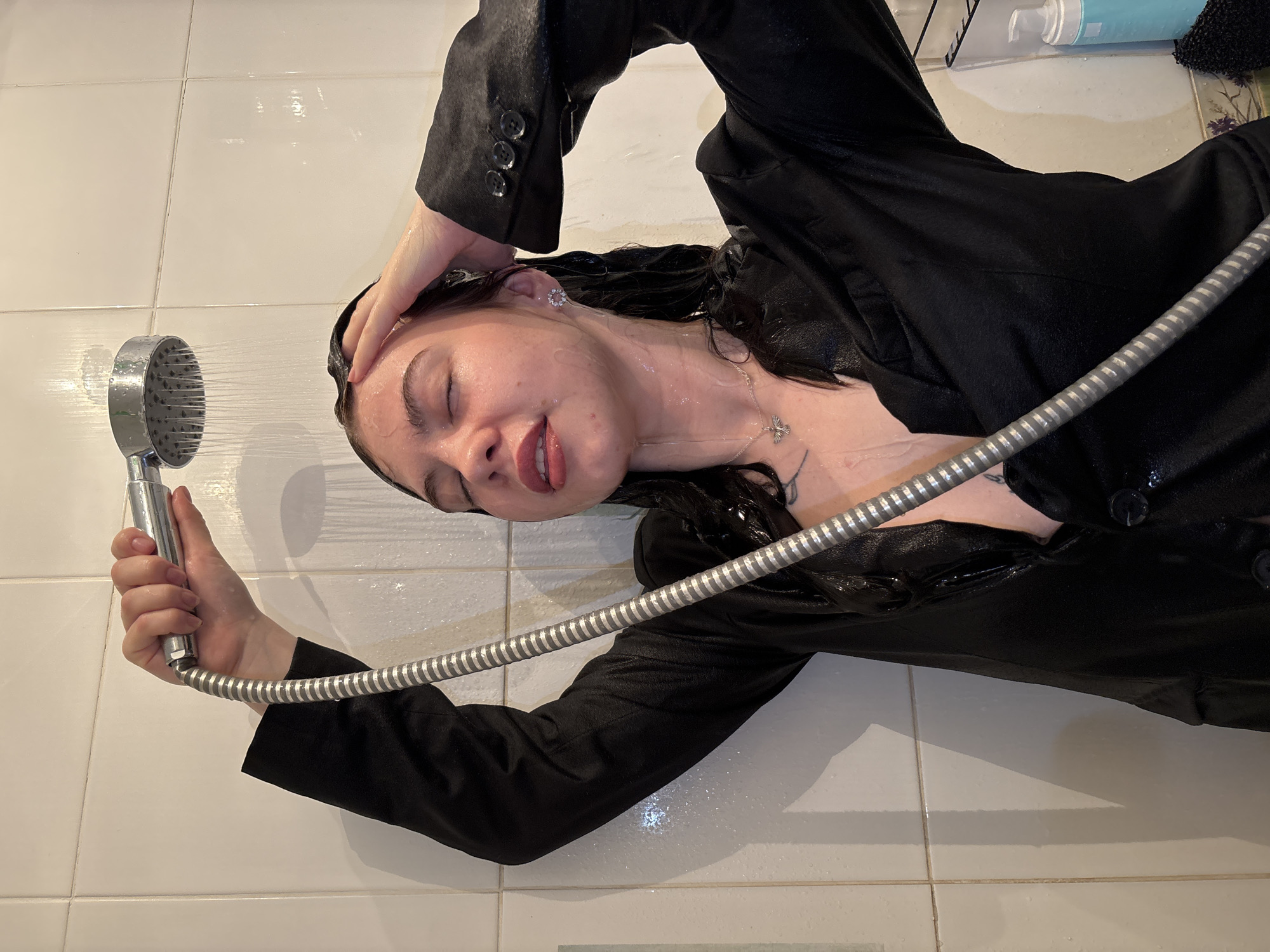 A woman in a wet black blazer lies on white tiled bathroom floor under a running shower. She holds the shower head with her right hand and rests her left hand on her forehead. Water is running over her face and hair. The photo was taken in Perm, Russia.