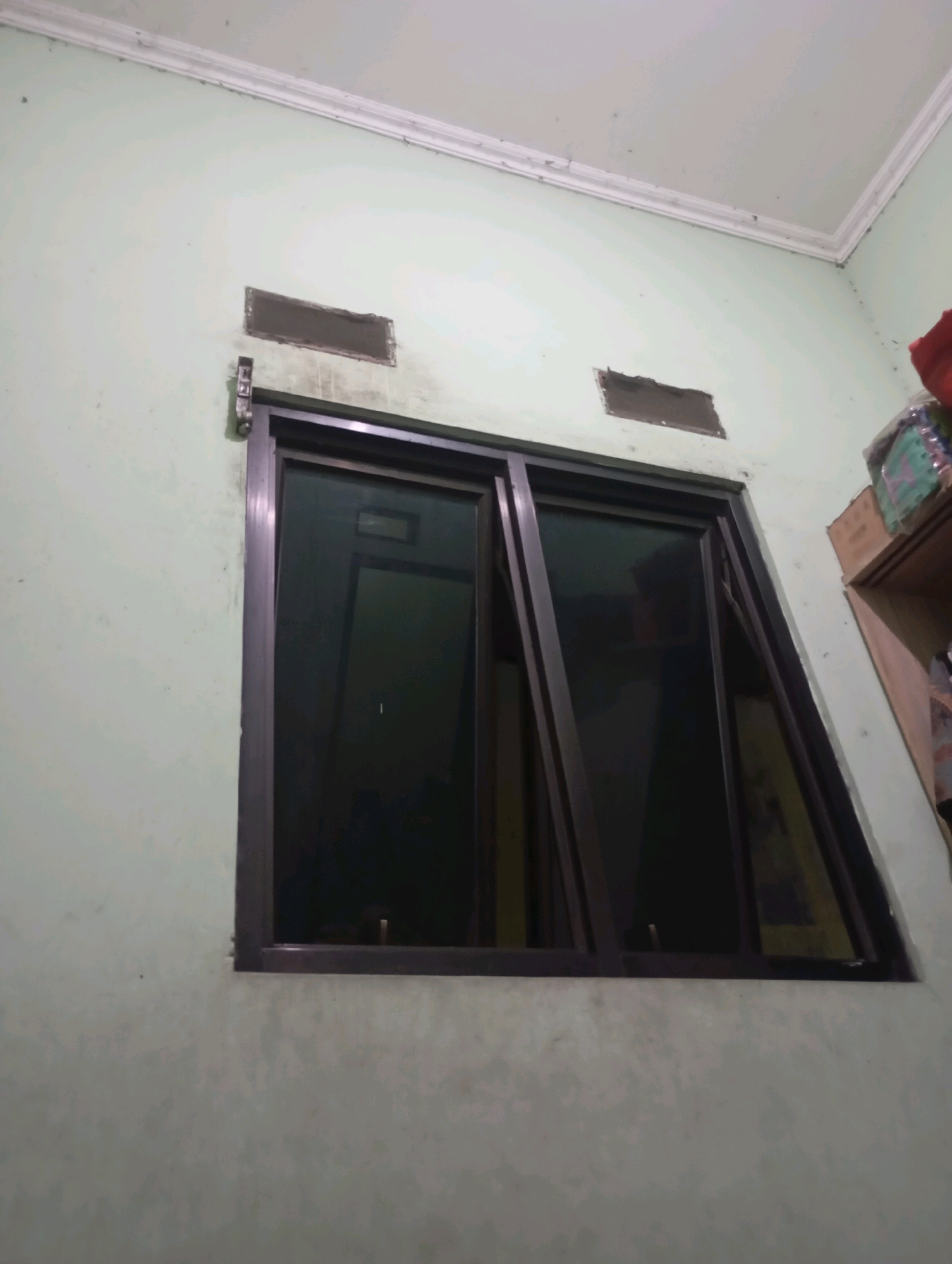 The image depicts a window with dark-tinted glass set into a light green wall. The window has a dark brown frame and appears to be a sliding or casement style, with one pane tilted open. Above the window, there are two rectangular vents integrated into the wall. The ceiling is white with molding. To the right of the window, a shelf is visible, holding various items, including boxes and what appears to be fabric. The wall shows some signs of wear and tear, with smudges and marks. The scene is indoors, likely in a residential or commercial building. The location is identified as Babelan, Indonesia.
