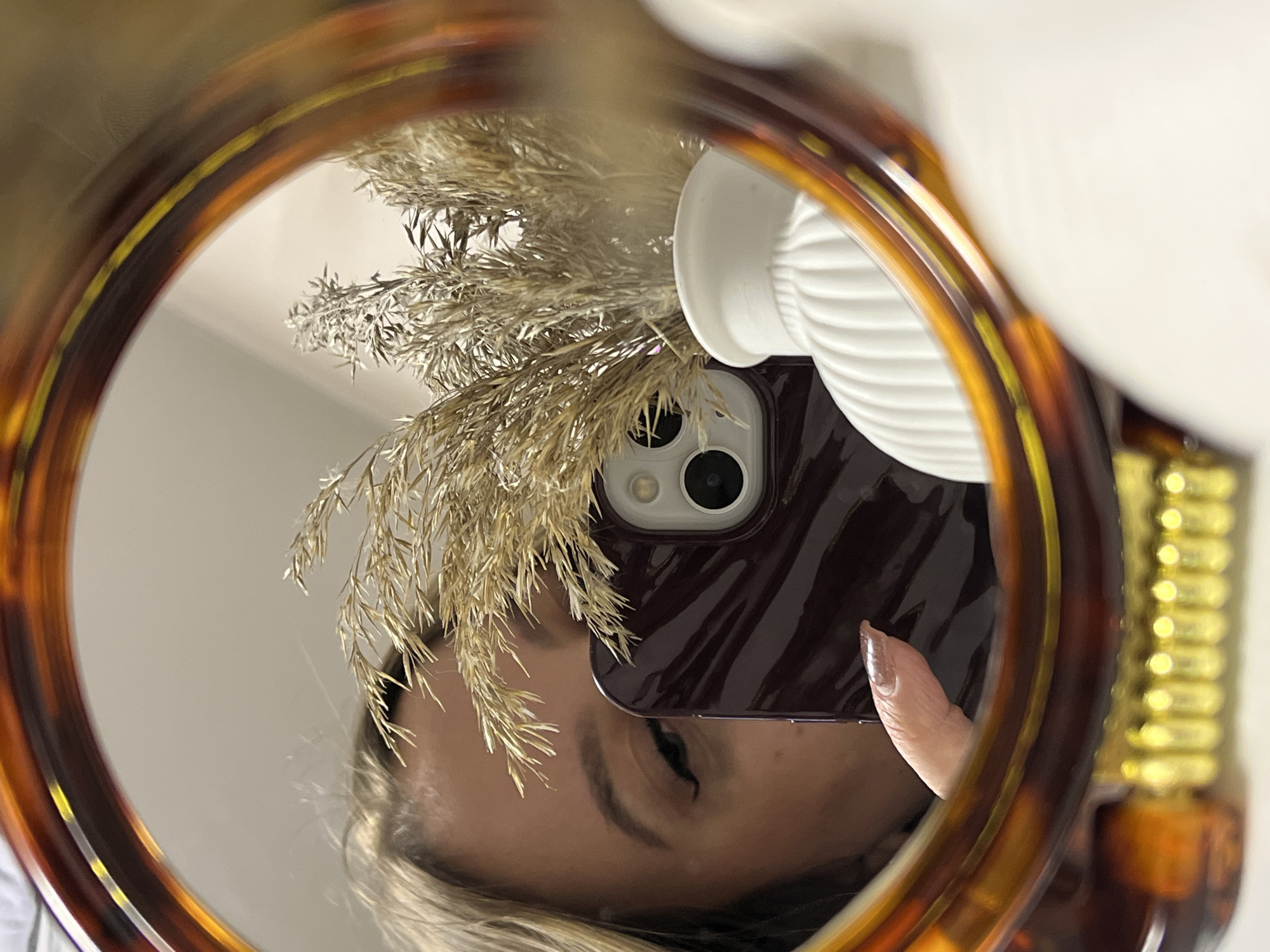 A mirror selfie, taken in Krasnodar, Russia, shows the reflection of a person and their surroundings. The image is framed by a round mirror with a distinctive tortoiseshell-patterned border. Reflected within the mirror, the lower part of a person's face is visible, featuring an eye with makeup and a sweep of light-colored hair. A smartphone, held by a hand with a manicured finger, occupies the center, its dark-colored case and dual-lens camera system clearly visible. It partially obscures the person's face. Behind the phone, a bundle of dried, feathery plants, resembling pampas grass, is arranged in a white, ribbed vase. A portion of another golden, ribbed decorative object is visible on the far right, also reflected in the mirror. The background reflection suggests a plain, light-colored indoor setting. The activity depicted is the act of taking a photograph of one's reflection. No text is visible in the image.