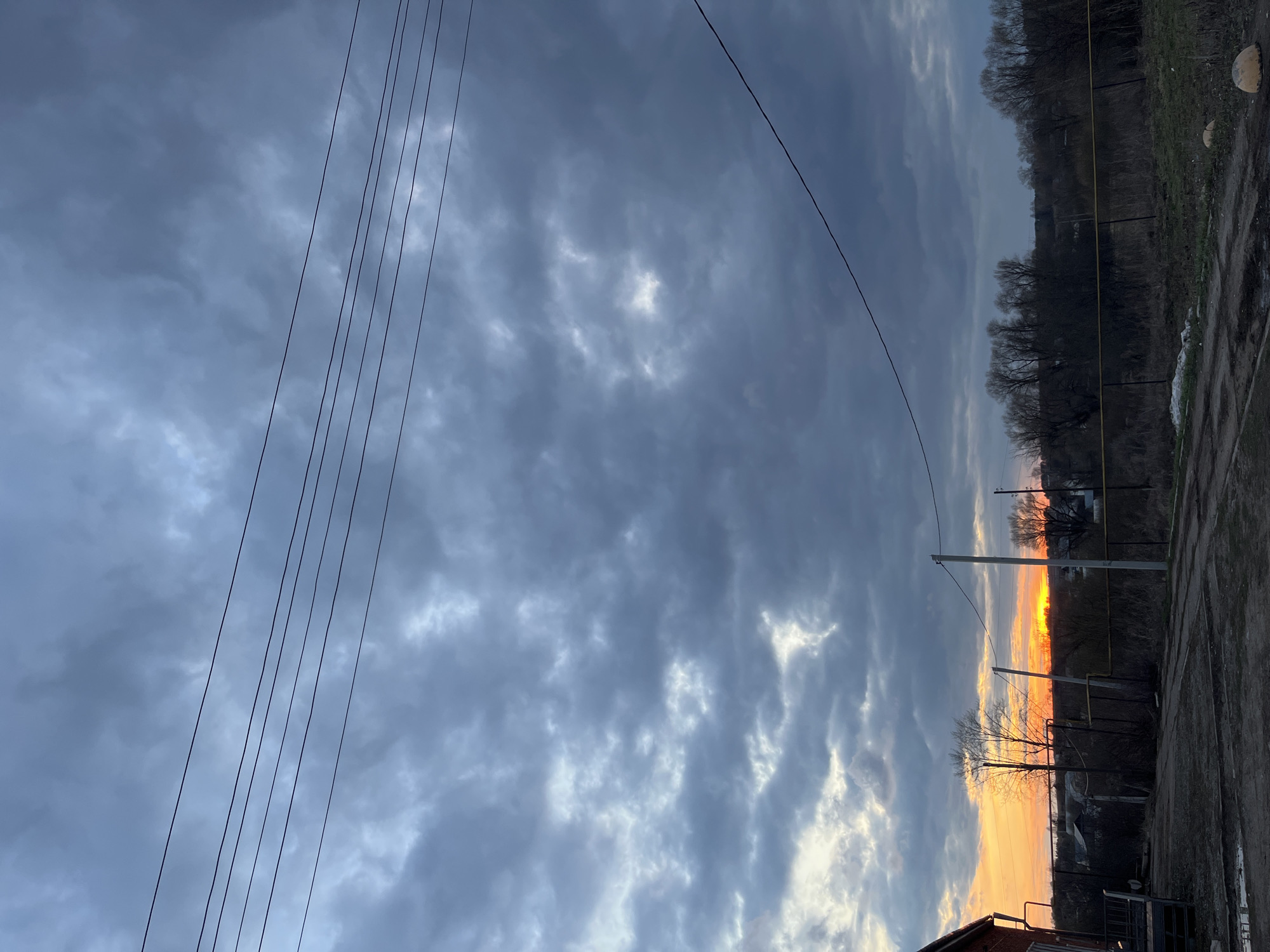 This image depicts a rural or semi-urban landscape in Кварса, Russia, captured during sunset. The sky is a dominant feature, filled with dramatic, dark grey clouds that show varying textures and densities. Patches of lighter clouds reveal the intense glow of the setting sun on the horizon, painting the lower sky with vibrant orange and yellow hues.
In the foreground, multiple dark utility lines or power cables stretch diagonally across the upper left and mid-right portions of the frame. These lines are supported by dark utility poles, one of which prominently features a vertical yellow gas pipe. The ground level in the immediate foreground is unpaved, appearing as dirt or uneven terrain. A small section of a building, possibly brick, is visible at the very bottom left corner.
The midground is characterized by a dense line of bare, leafless trees, silhouetted against the bright sunset and the overcast sky, suggesting either late autumn, winter, or early spring. A fence line or a series of dark posts runs through this area. In the distance, a dark, tree-covered slope or hillside forms the backdrop, beneath the dramatic cloud cover. The strong contrast between the radiant horizon and the dark foreground elements, set against the cloudy sky, creates a captivating and slightly melancholic atmosphere. No visible text is present in the image.