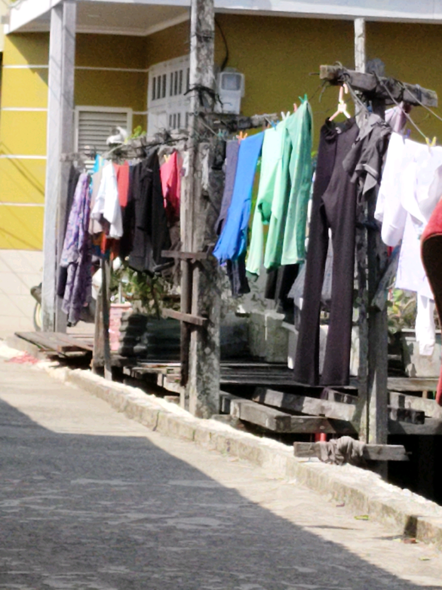 The image depicts a clothesline with various garments hanging to dry outside a building in Balikpapan, Indonesia. The building has a bright yellow exterior and white trim. The clothesline is supported by wooden posts, one of which appears to be concrete. Garments include shirts, pants, and what looks like a patterned dress or skirt. Some clothes are pinned to the line with clothespins. The ground in front of the building is a concrete path, and there is a body of water visible in the background behind the clothesline. The scene suggests a residential or domestic setting.