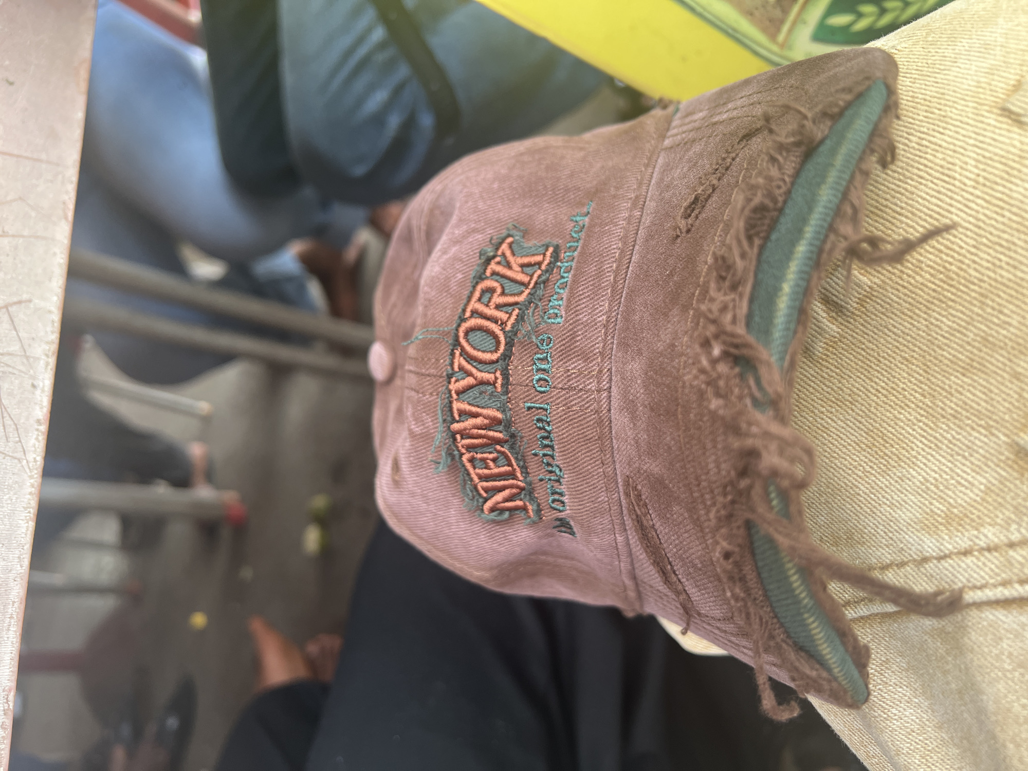 An overhead, angled shot captures a worn brown baseball cap embroidered with "NEW YORK" in two-tone lettering (dark green outline, orange fill), resting atop a person's lap. The cap's brim is frayed and tattered, revealing the green lining. The person's clothing includes what appears to be tan denim on the right side of the frame and a dark fabric (possibly black pants) beneath the cap. The background is indistinct, but there's evidence of what appears to be interior space, including metal frame chairs/seats and glimpses of other people's legs and feet. Based on the fact the hat is a New York themed hat, and the requested location is Aso Nigeria, there is not sufficient information to conclude where this picture was taken, only where it may be located based on the information provided.