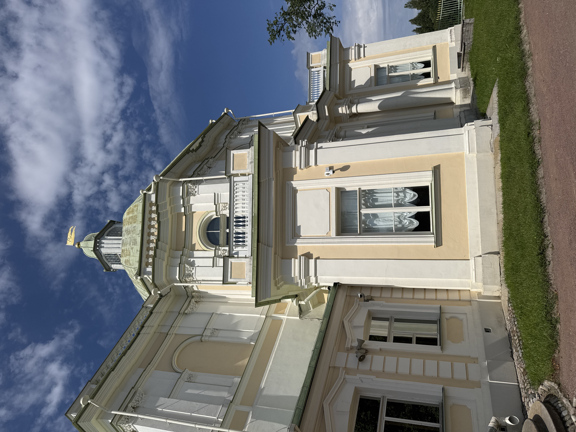 The image captures a low-angle view of a light yellow and white baroque-style building, likely a palace or grand residence, under a bright blue sky with scattered white clouds. The architecture features ornate moldings, columns, arched windows, and a green dome topped with a golden flag bearing some inscription. The building is surrounded by lush green grass and a paved path. The scene is devoid of any people. The overall impression is one of stately grandeur and historical architecture. This building is likely part of the Oranienbaum palace complex in Lomonosov, Russia, given its architectural style and the context of similar structures in the area.