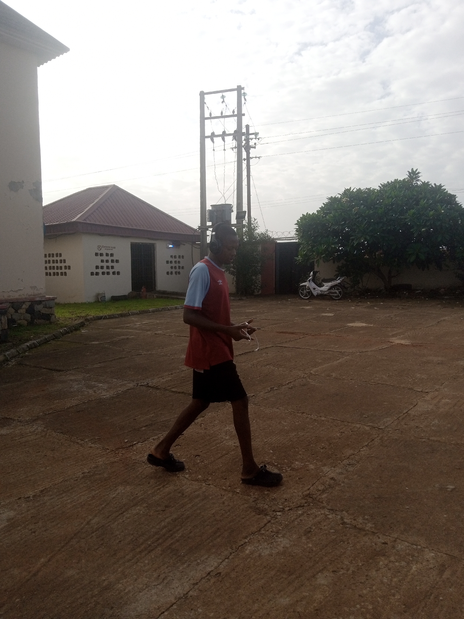 In Jos, Nigeria, a young man wearing a red and blue shirt and black shorts walks across a paved courtyard. He is holding a white object in his hands and appears to be looking at it. In the background, a small building with a red roof is visible, as are power lines and a tree. A white motorcycle is parked near the tree. The sky is overcast.