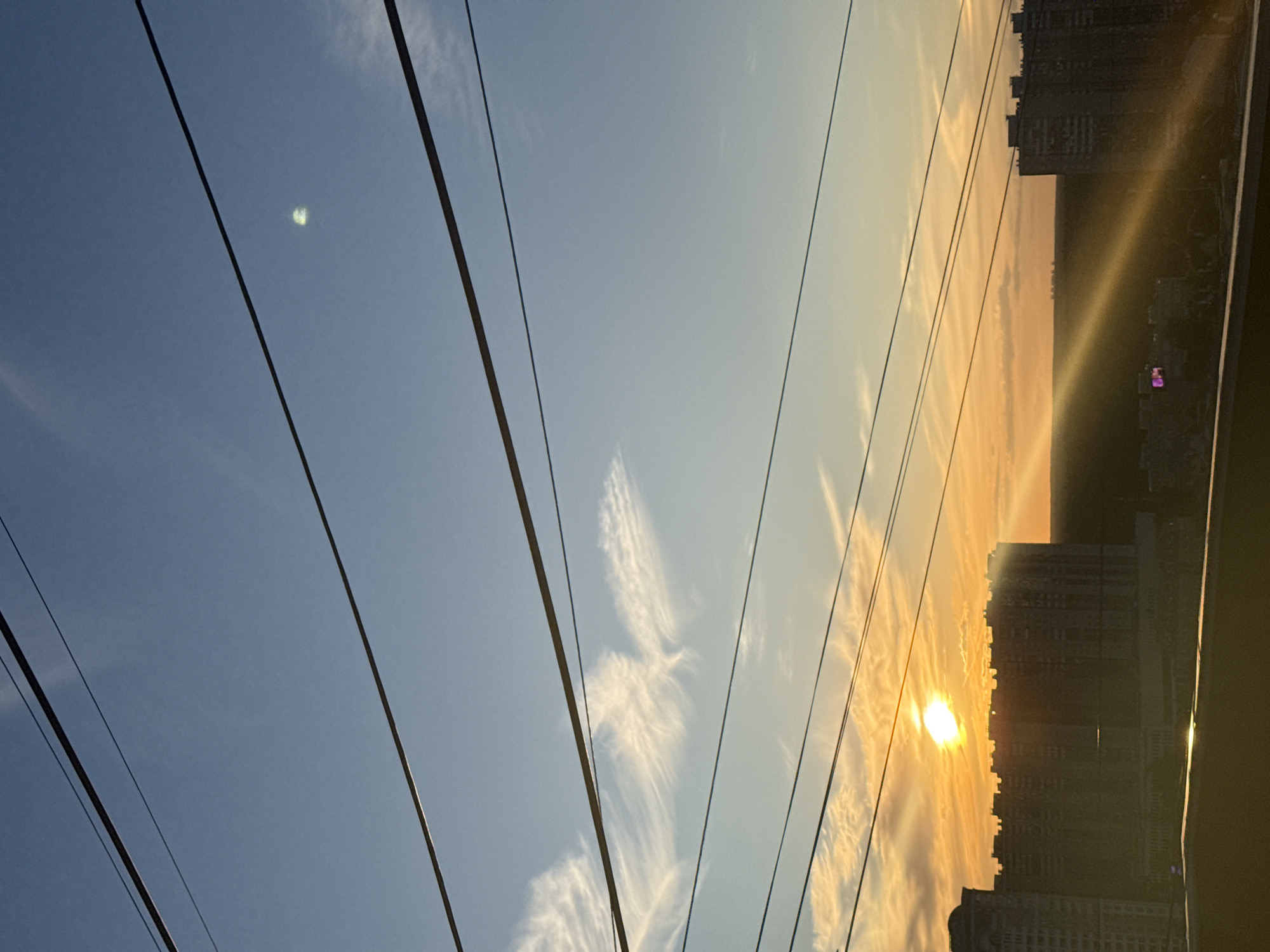 This is a photograph taken from a low angle, looking up towards a clear blue sky with wispy clouds. Several power lines stretch diagonally across the frame, dominating the foreground. In the distance, silhouetted buildings rise against a vibrant sunset. The sun is low on the horizon, casting a warm orange and yellow glow across the sky and the clouds, creating a dramatic effect. The buildings appear to be apartment complexes, with multiple windows visible. The overall scene suggests an urban environment at dusk. The sun's glare creates lens flares and bright streaks of light across the image, particularly over the buildings. The sky transitions from a deep blue on the left to a pale yellow and orange on the right, indicating the setting sun. The clouds are thin and feathery, catching the sunlight and appearing brightly illuminated. The image appears to be taken outdoors, possibly from a window or balcony, given the perspective and the presence of the power lines. There is no visible text in the image.