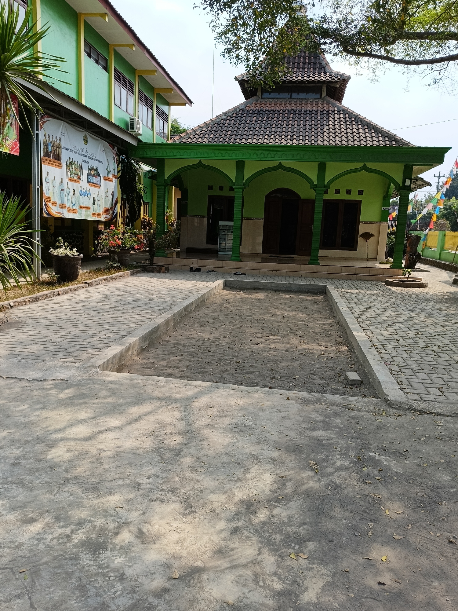 The image shows a small mosque in Barukan, Indonesia, situated next to a green school building. The mosque features a traditional tiered roof and a green facade with arched doorways. Paved walkways made of interlocking bricks lead to the mosque, and a sandy area is visible in the foreground. The surrounding environment includes trees, potted plants, and a yellow fence in the distance. The sky is clear and bright, suggesting a sunny day. There are no people visible in the image.
