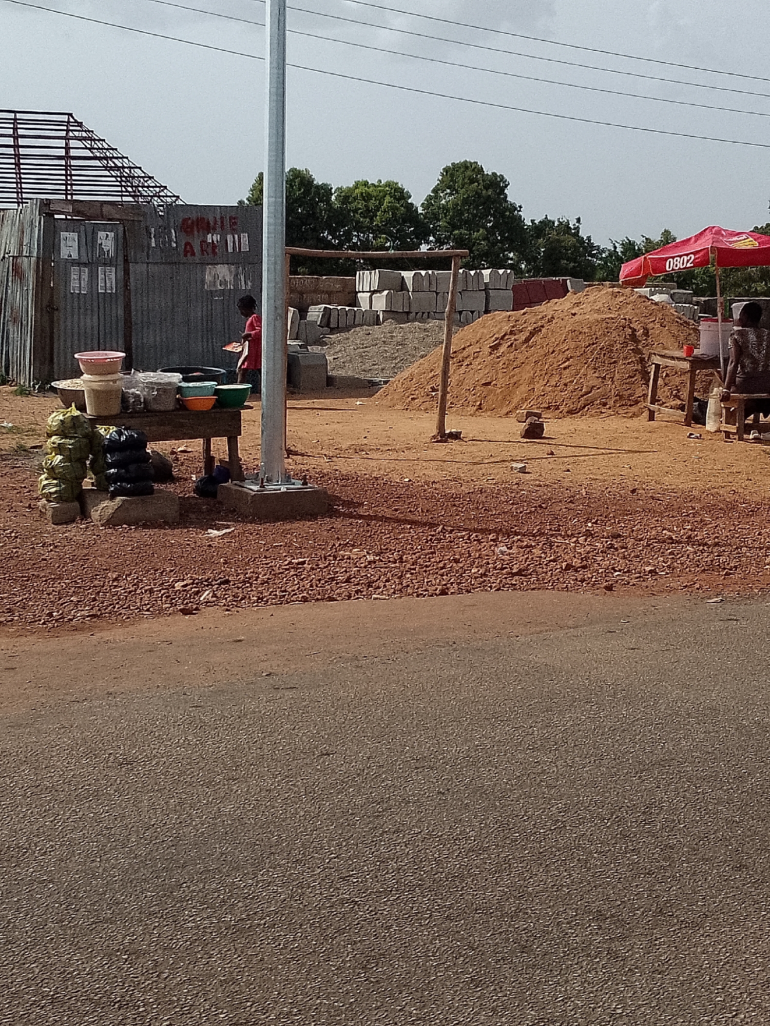 The image depicts a roadside scene in Kuru Karama, Nigeria. There's a corrugated metal structure with a partially built roof, and a sign with faded writing. A person wearing red clothing is visible near a table laden with bowls and packages. To the right of the table stands a utility pole. Behind this area, a pile of sand and stacks of grey concrete blocks are observed. Further to the right, under a red umbrella bearing the number 0802, sits an individual on a wooden bench near a table and various containers. The ground in the foreground is reddish-brown gravel, and a portion of a paved road is at the bottom of the frame. Overhead power lines stretch across the top of the image. The sky is overcast.