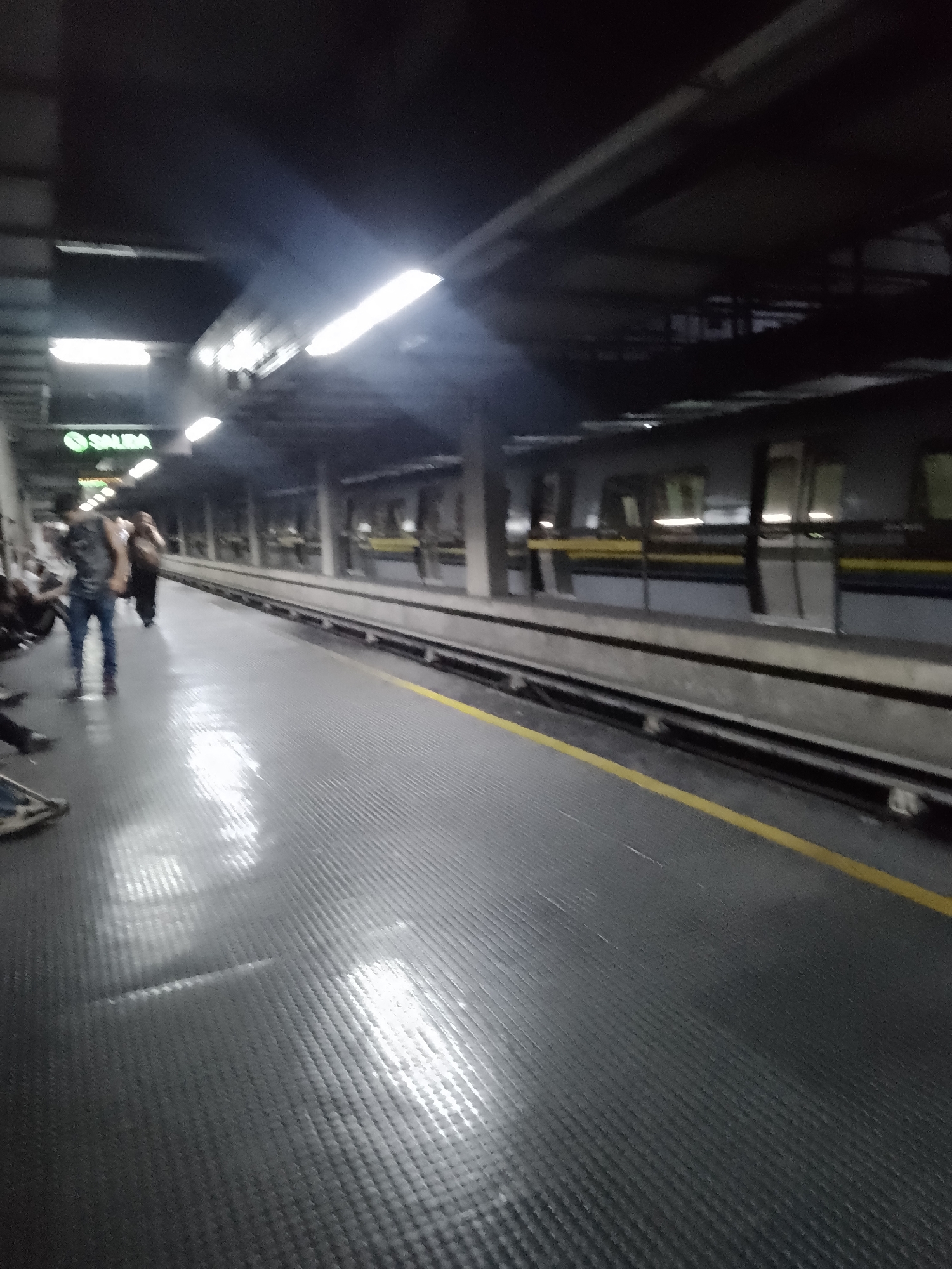 A ground-level, slightly blurred image shows the interior of a subway station in Caracas, Venezuela. The platform is visible, with a gray tiled floor reflecting light. A yellow safety line runs along the edge of the platform. A subway train is parked at the platform. The train is light gray with dark gray and yellow accents. Several people are visible on the platform. There is a green sign labeled "Salida," which translates to "Exit." Overhead lighting illuminates the platform and the train. The ceiling appears to be a dark gray structure.