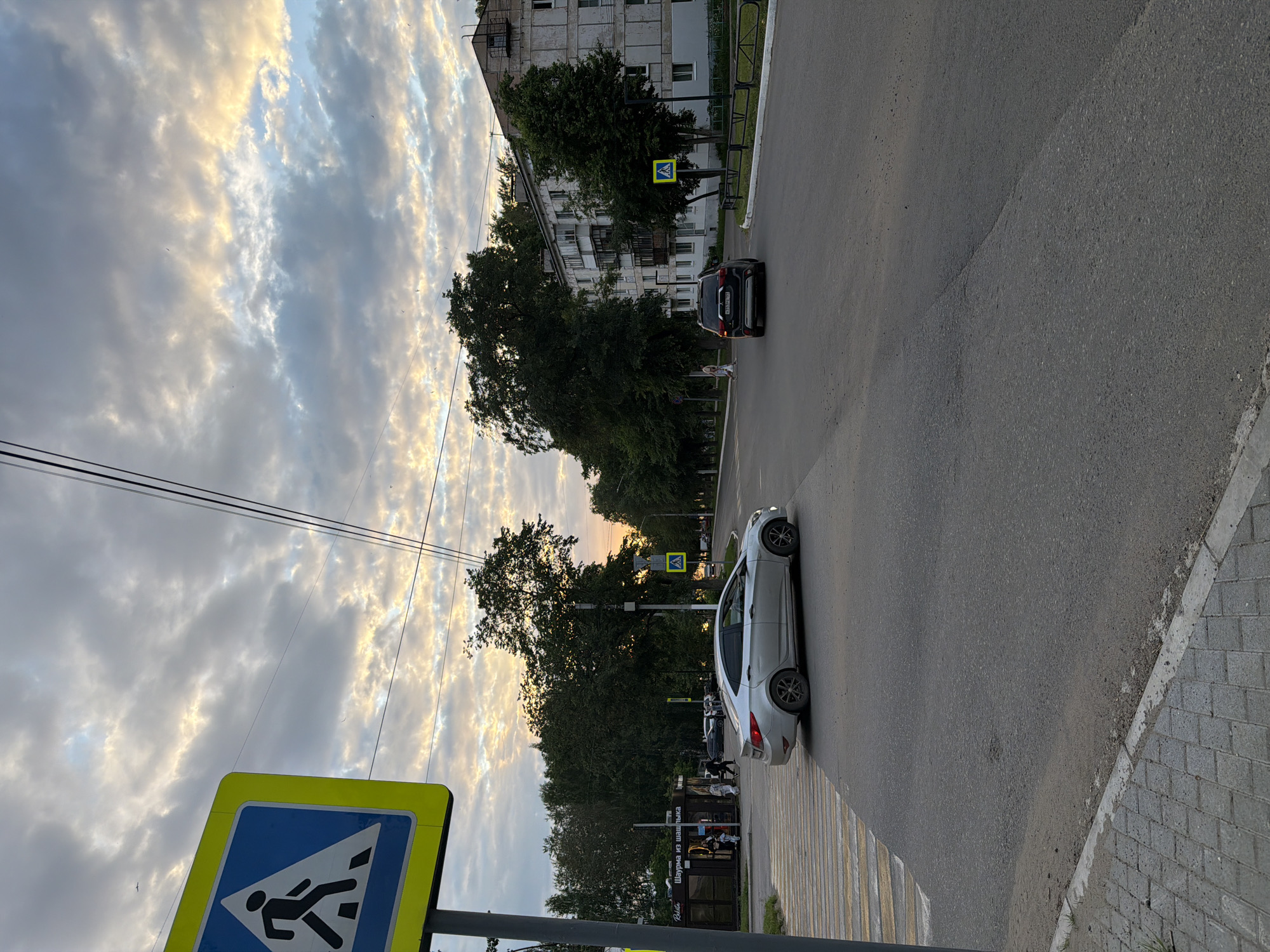 The image depicts a crosswalk in the city of Chaikovsky, Russia, with a silver car stopped at the intersection. The sky is cloudy with patches of sunlight breaking through, indicating either sunrise or sunset. A pedestrian crossing sign is visible on the left side of the road, and another similar sign is located further down the street on the right. There are trees lining the street and a multi-story apartment building in the background. A few people are visible walking near a small kiosk on the left side of the road. The road surface appears to be asphalt.