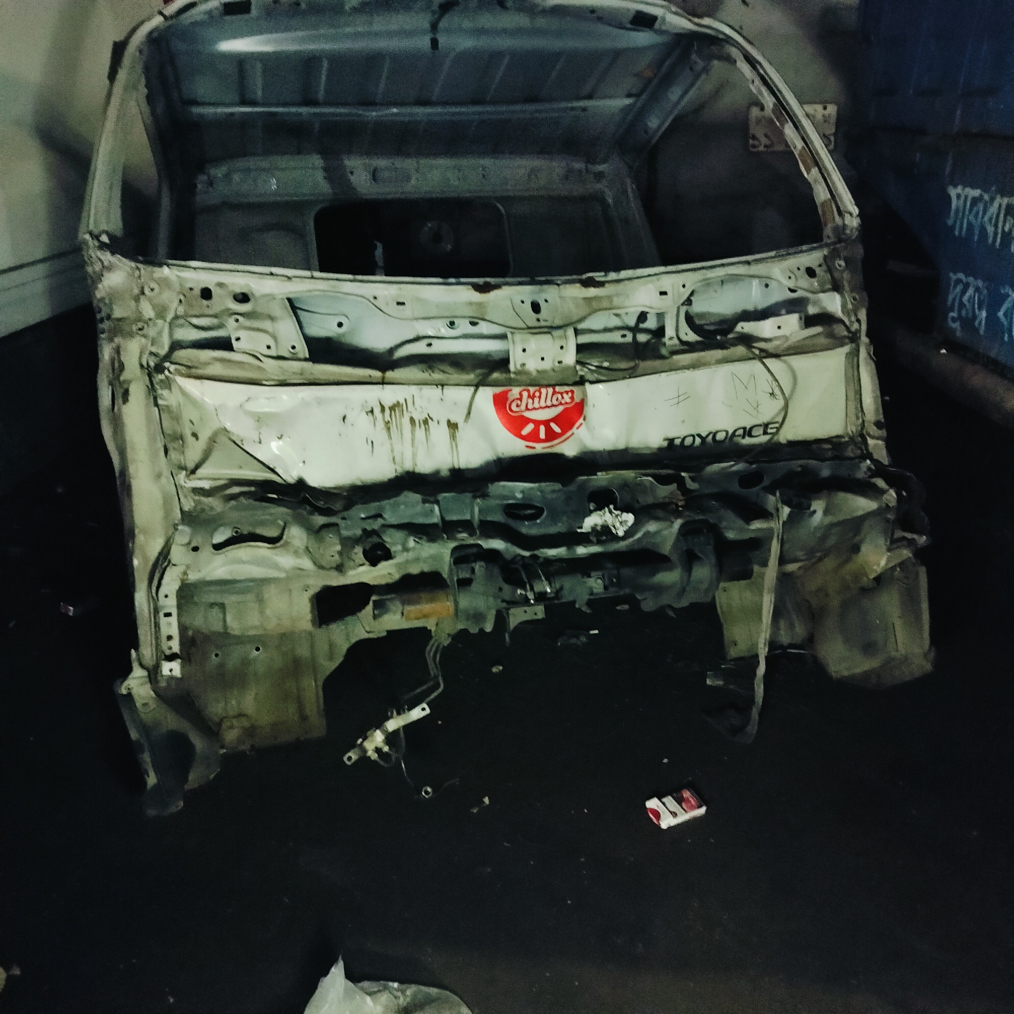 The image shows the damaged cab of a white TOYOACE truck, marked with a "Chillox" logo. It is devoid of doors, windows, and the front portion, revealing a hollowed interior. Scrawled writing is visible on the body. The vehicle remains are in a dark, confined space, possibly a garage or storage area. Debris and discarded objects lie on the ground near the cab. The location is indicated to be in an area of Unknown, Unknown.