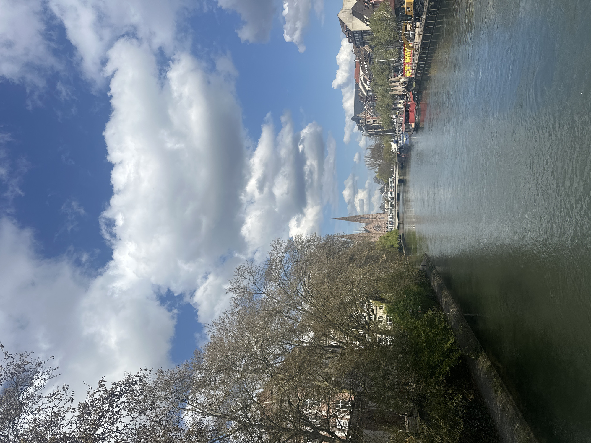 This image captures a view in Strasbourg, France, along the Ill River. A church steeple is visible above trees, likely belonging to the Église Saint-Paul. The river is flanked by buildings and trees on both sides. Along the opposite bank, there are moored boats. The sky is bright with puffy white clouds. The trees in the foreground appear bare, suggesting either late autumn, winter, or early spring. No people are clearly visible in the scene.