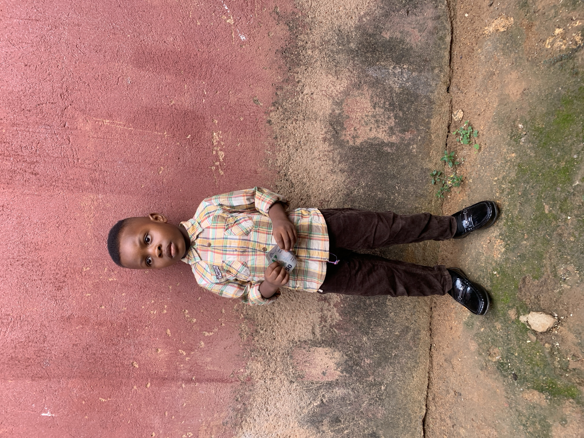A young boy lies on the ground, oriented lengthwise on the right side of the frame, on a paved surface alongside a wall painted maroon. The boy is dressed in a patterned button-down shirt, brown pants, and shiny black loafers. He is holding a small paper currency in his hands. The paved surface exhibits a color gradient from light brown to green with moss and dirt. A crack runs vertically through the pavement, separating the boy from the wall. Given the visual details, the image is likely taken outdoors in a residential area, possibly in Jos, Nigeria.