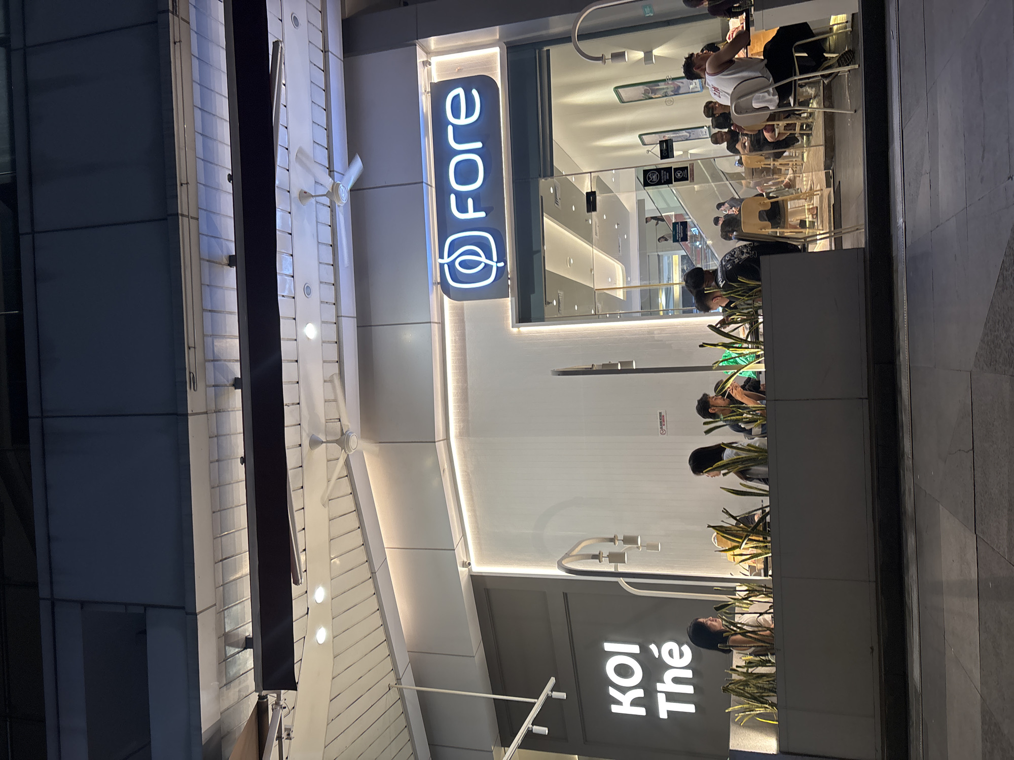 This image captures the exterior of a modern cafe, "KOI Thé," and an adjacent establishment with a neon sign that reads "fore." The scene is set at night in a public space, possibly a mall or promenade, in North Jakarta, Indonesia.
In the foreground, a low planter box filled with tall, spiky green plants separates the cafe's outdoor seating area from the pedestrian walkway. Several individuals are seated at tables within this outdoor space, some facing away from the camera, engaged in conversation. The lighting is a mix of warm indoor illumination spilling from the cafe and the cooler glow of the "fore" sign.
The "KOI Thé" signage is prominently displayed in white neon letters on a dark grey background. Adjacent to it, the entrance to the "fore" establishment is visible through large glass panels, revealing more seating and patrons inside. The interior lighting of "fore" is bright and contemporary, highlighting sleek furniture and a minimalist aesthetic. Two ceiling fans are visible in the upper section of the frame, indicating a warm climate. The overall impression is one of a lively evening scene at a commercial establishment in a developed urban area.
