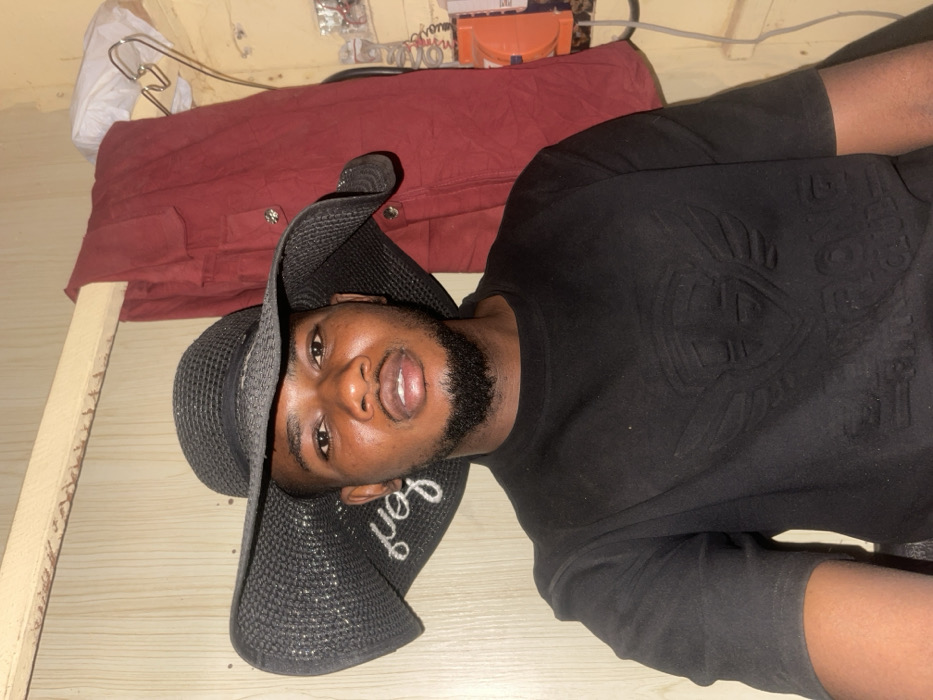 A man is lying on a wooden surface. He wears a black t-shirt with an embossed design and a black sun hat. The hat has a word embroidered on its brim. Behind him is a red garment or cloth piece, possibly on a bed. In the background, miscellaneous items are visible, including some white plastic bags and electrical wiring. The man appears to be posing, looking directly at the camera.