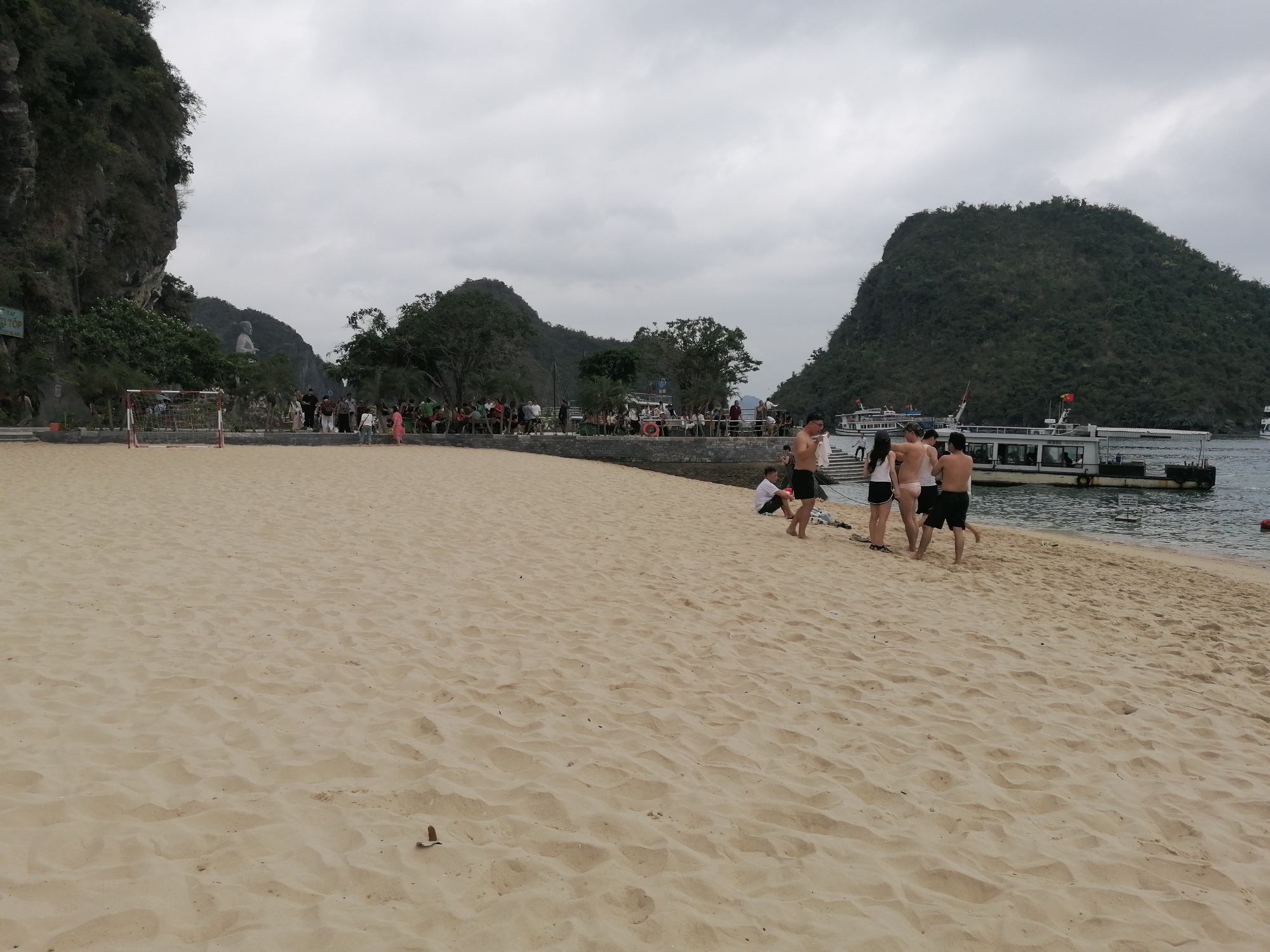 The image presents a vibrant beach scene at Ti Top Island in Ha Long Bay, Vietnam, under an overcast sky. In the foreground, a wide expanse of light-colored sandy beach dominates, showing numerous footprints and undulations from recent activity. Several individuals, some in swimwear and others in casual clothes, are visible near the water's edge on the right, either walking towards or away from the calm, dark water where a few boats are docked.
Behind the beach, a paved promenade runs along the base of a lush, tree-lined embankment. A small, red and white soccer goal is positioned on the left side of this paved area, hinting at recreational use. Along the promenade, groups of people are scattered, some seated or gathered, while others walk. A partially visible sign on the far left clearly displays "ĐẢO TI TỐP," which translates to "Ti Top Island," confirming the specific location.
In the middle ground, several passenger boats or tour vessels are moored or docked, one of which prominently flies the red and yellow Vietnamese flag. The background is characterized by the iconic, verdant karst mountains and islets typical of Ha Long Bay. A distinctive large white statue of a person is visible on a distant, tree-covered hill, overlooking the bay. The sky is uniformly gray and cloudy, indicating overcast weather throughout the daytime scene, providing a soft, diffused light across the landscape and the water. The overall impression is of a popular tourist destination where visitors enjoy the beach and boat tours amidst the stunning natural scenery.