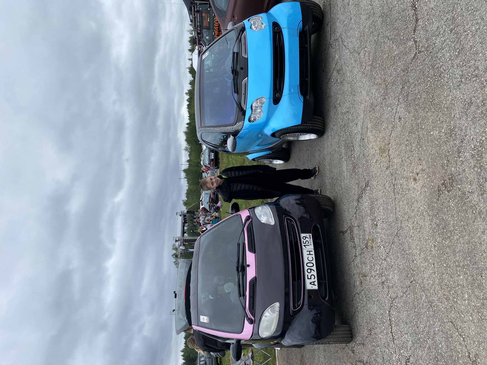 An outdoor photograph shows two smart cars parked side-by-side on an asphalt lot. The car on the left is black with a pink accent on the hood and trim, and the car on the right is bright blue with a black top and silver rims. A young person in a black puffer jacket and black pants stands between the two cars, looking at the camera. In the background, a stage with lights and a crowd of people are visible, suggesting an event or festival. Trees line the horizon under a cloudy sky. The license plate of the black car reads "A590CH 159 RUS", indicating it is registered in the Perm Krai region of Russia, where the city of Perm is located.