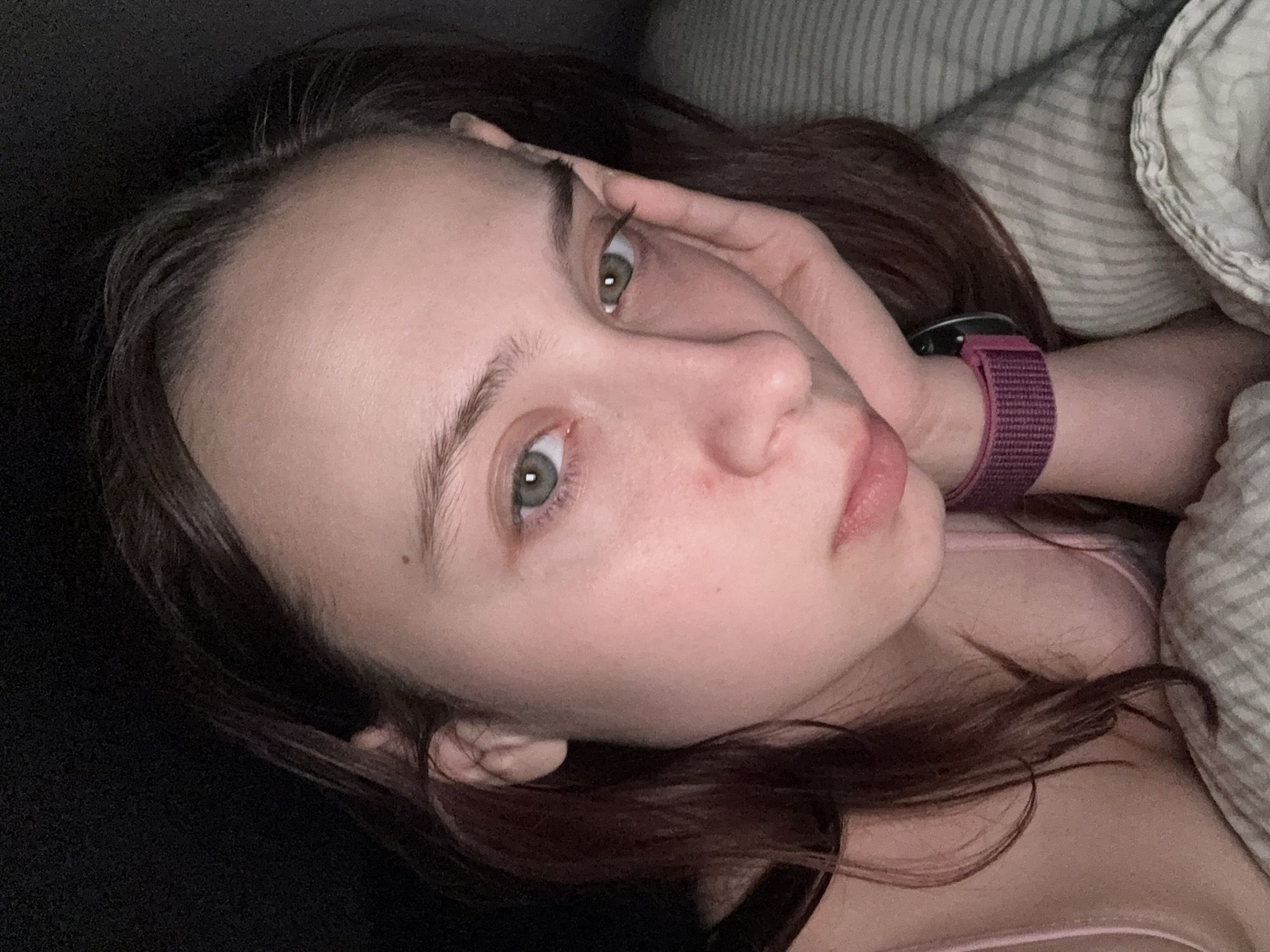 A young woman with light skin, green eyes, and dark brown hair lies in what appears to be a bed. She is resting her head on her hand, with a pink smartwatch visible on her wrist. Bedding and part of a headboard are also visible. The location is not discernible from the image.
