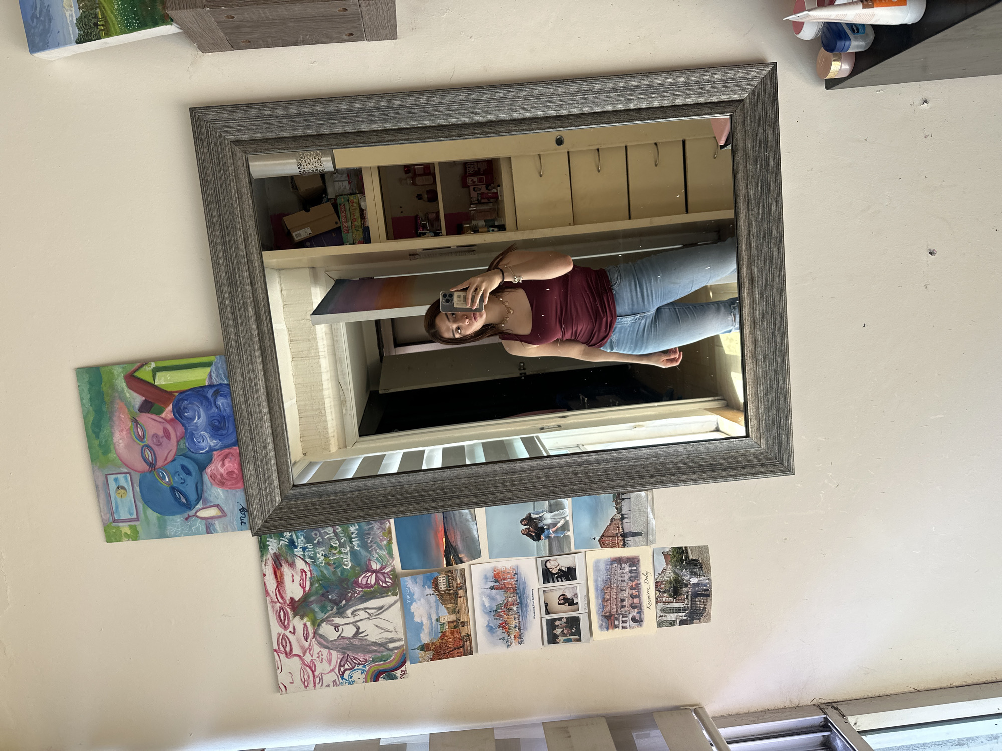 A woman takes a selfie in a mirror. The mirror hangs on a wall in front of a painting and postcards. The mirror reflects a room with shelving and cabinets. The exact location within Puebla, Mexico is impossible to ascertain from the provided information.